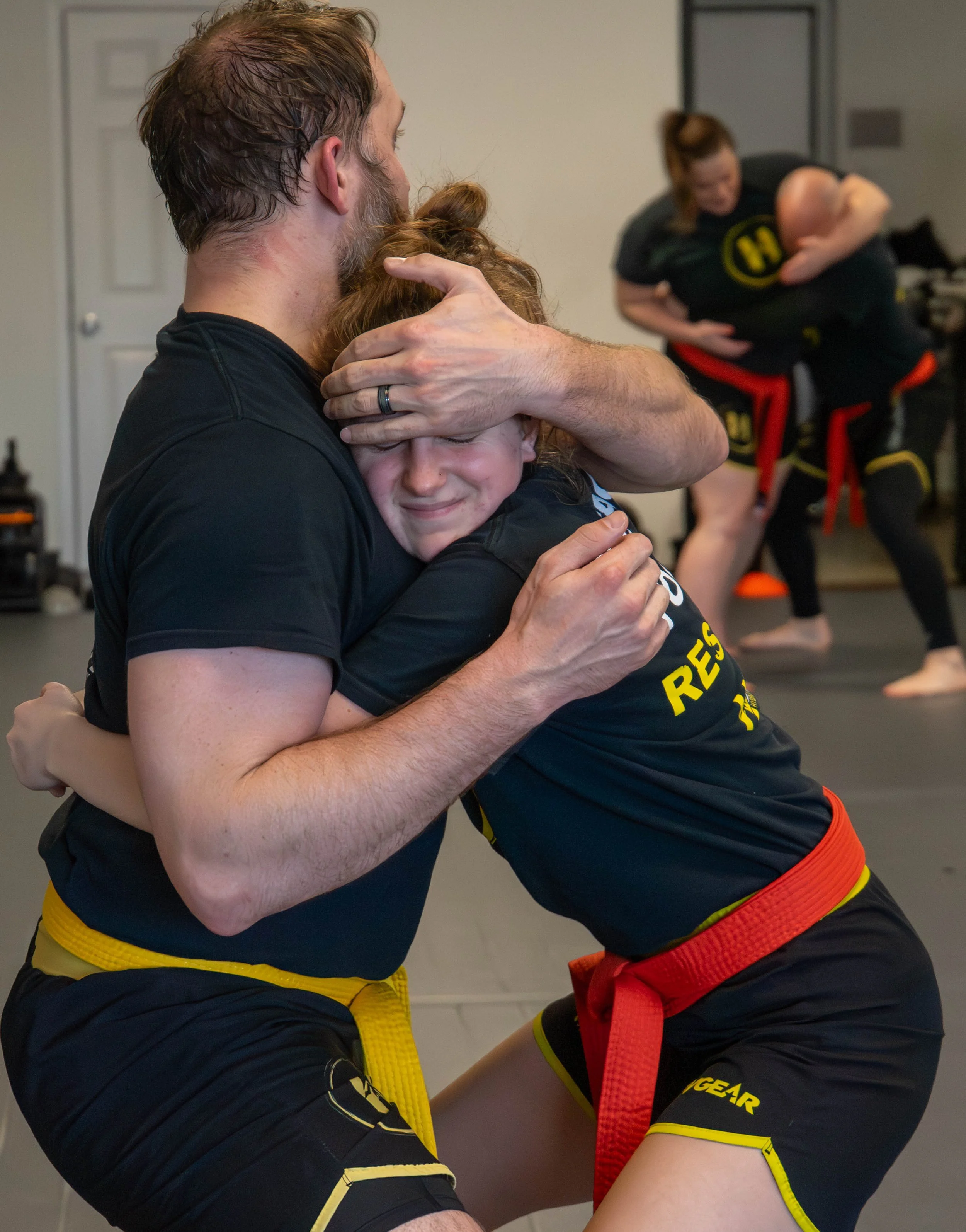 A man and a young girl practicing Brazilian Jiu-Jitsu. The man is holding the girl in a close embrace while she is crying and smiling. They are both wearing black martial arts uniforms with yellow and red belts. In the background, other women are practicing techniques.