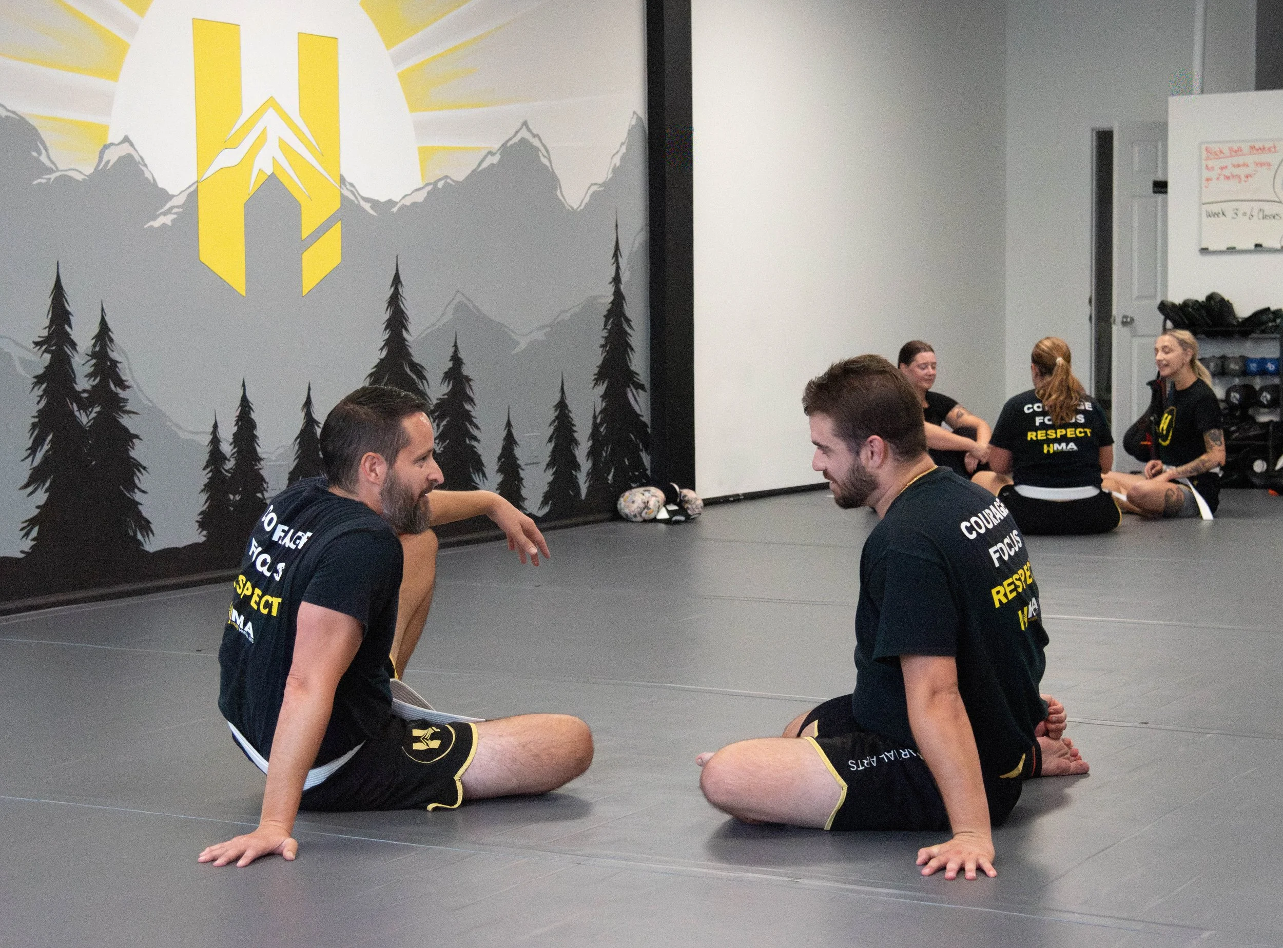 Martial arts students sitting on a gray mat, engaging in a conversation during class in a gym with mountain and forest mural on the wall.