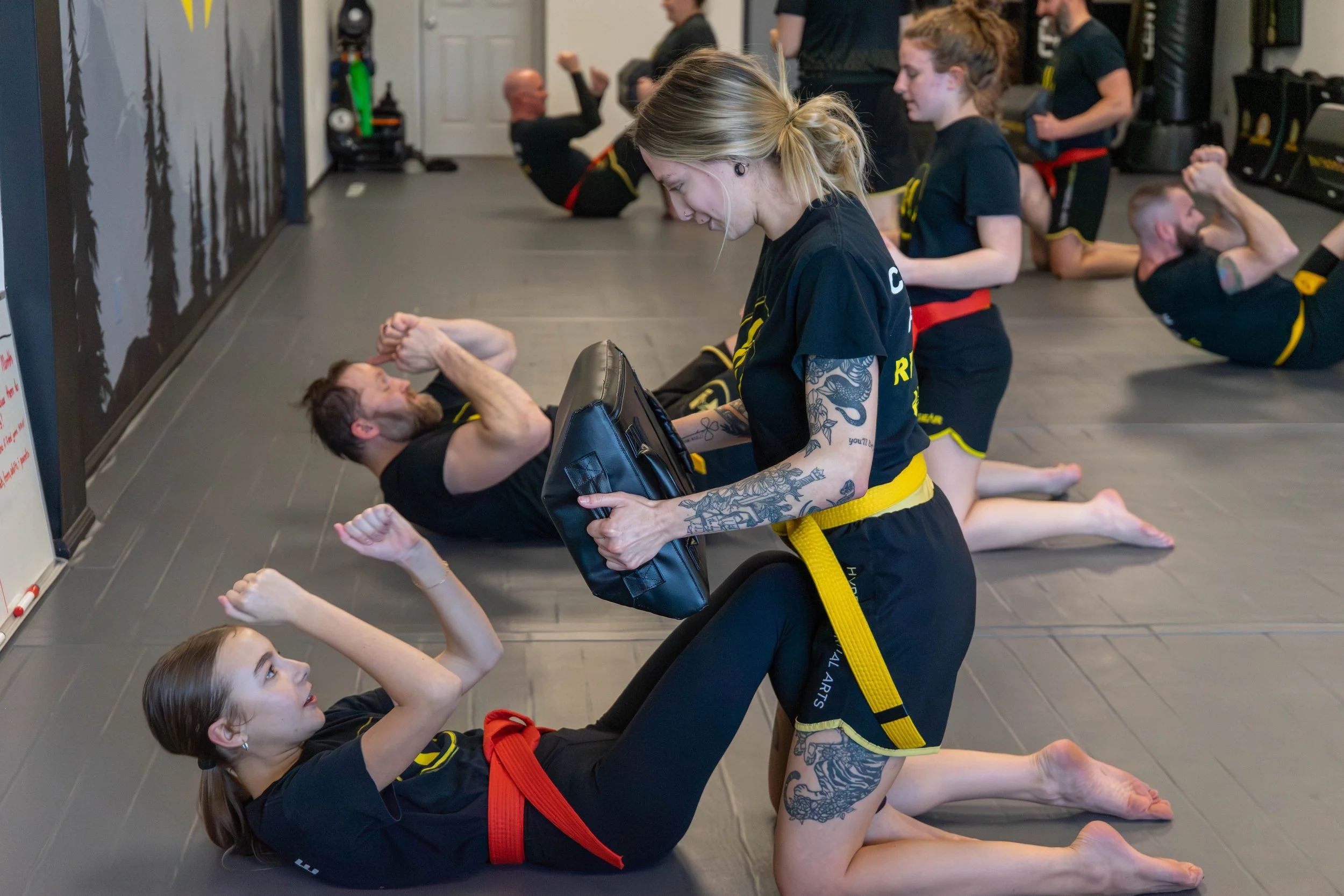 Martial arts class with students practicing techniques on mats, instructor in center holding a pad, other students in black uniforms with red or yellow belts, some stretching or in sparring positions.