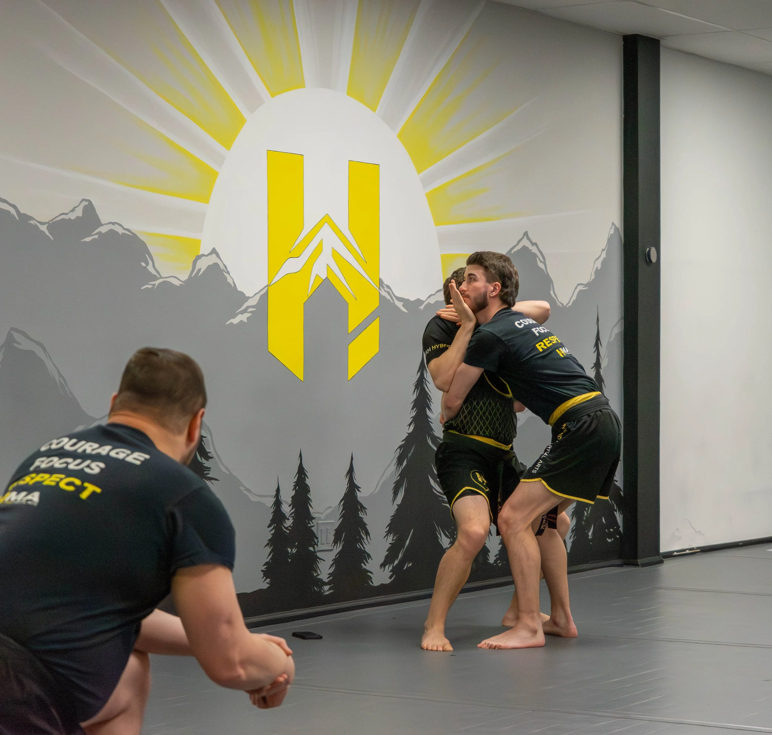 Two men practicing Brazilian Jiu-Jitsu on a black mat in a gym with mountain and trees mural and a large yellow and white logo on the wall. A third person watches nearby.