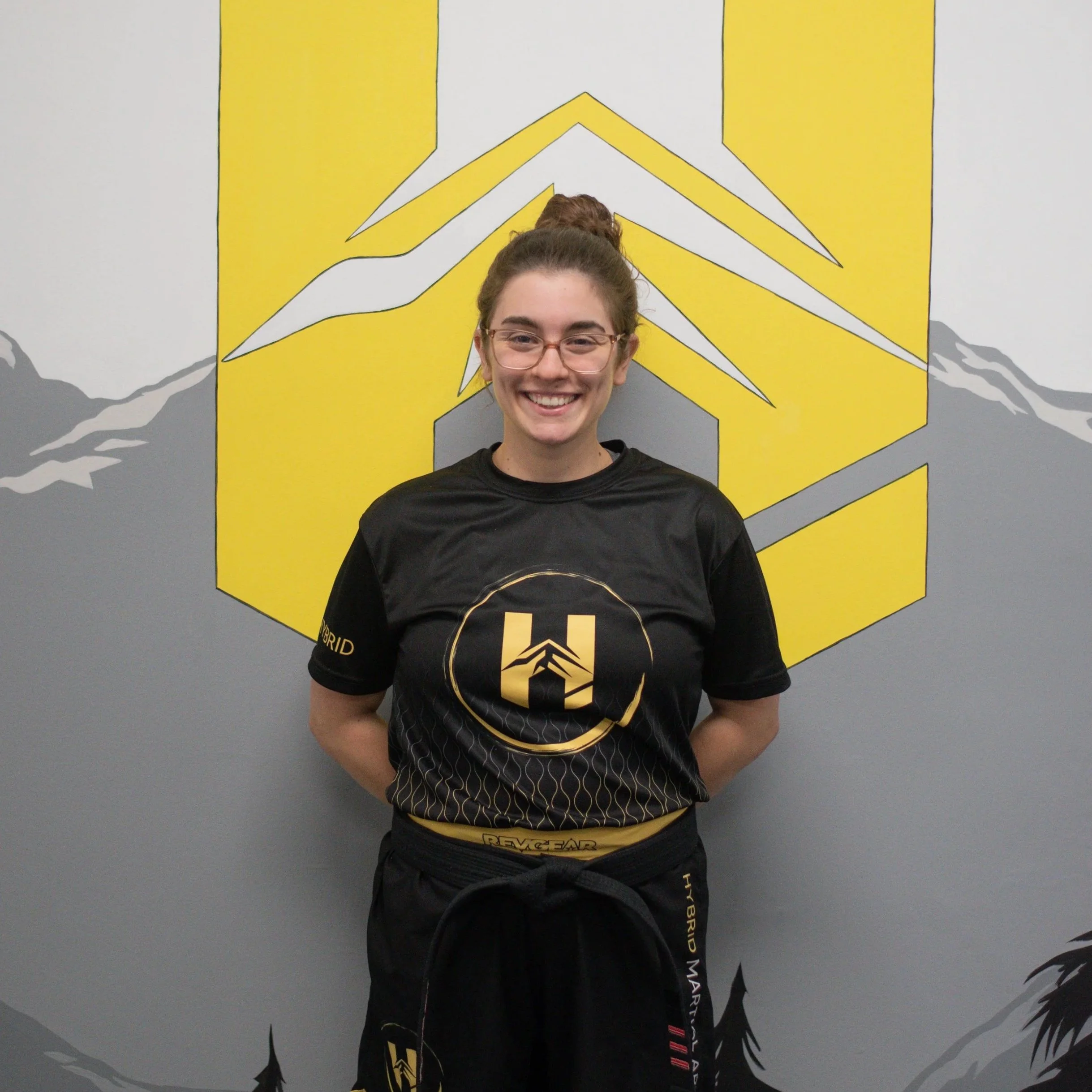 A young woman with glasses and a ponytail, smiling, wearing a black sports shirt with a yellow logo and black athletic pants, standing in front of a wall with mountain and yellow geometric graphics.