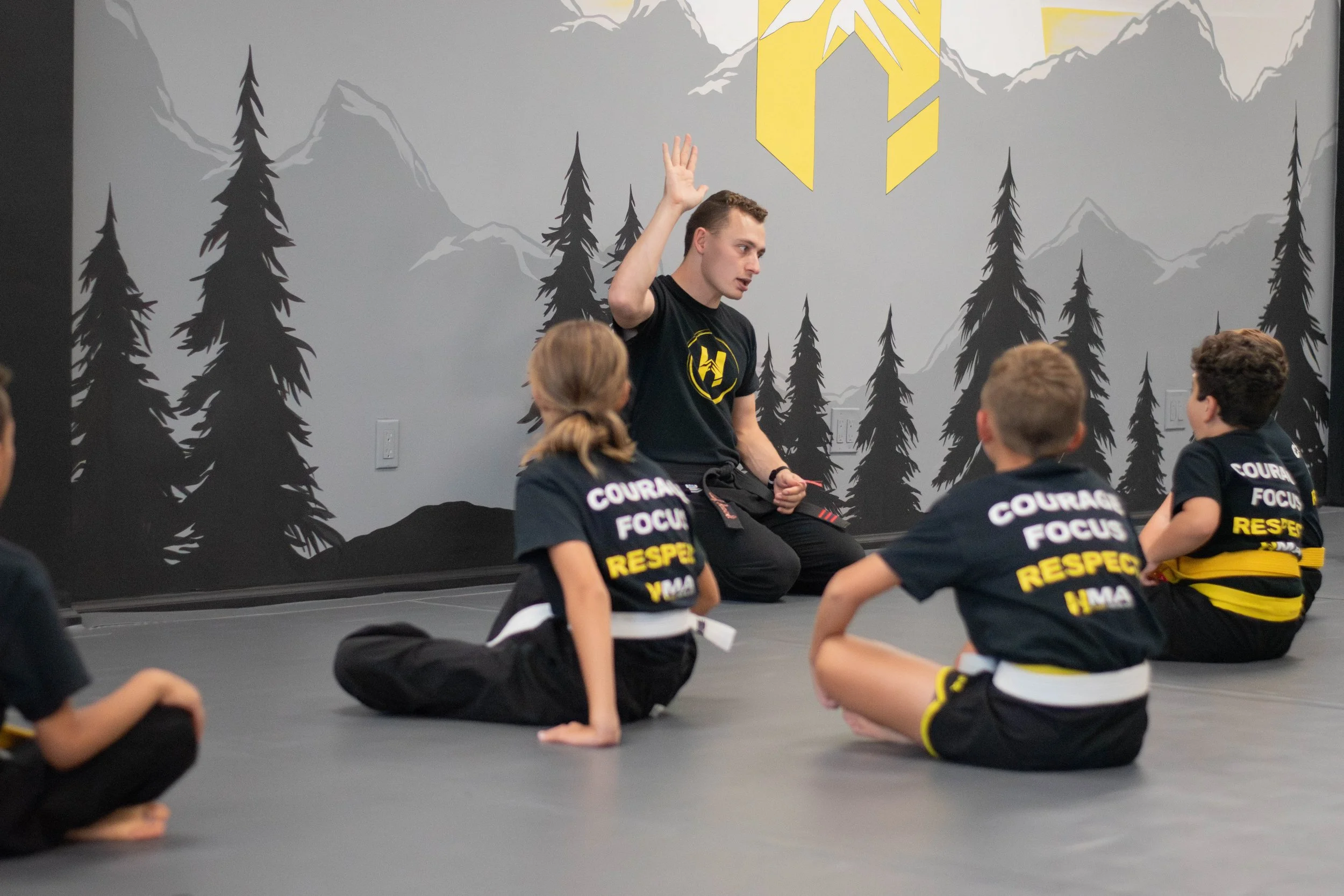 Martial arts instructor teaching a class of young children seated on the mat, with a mountain and forest mural background.