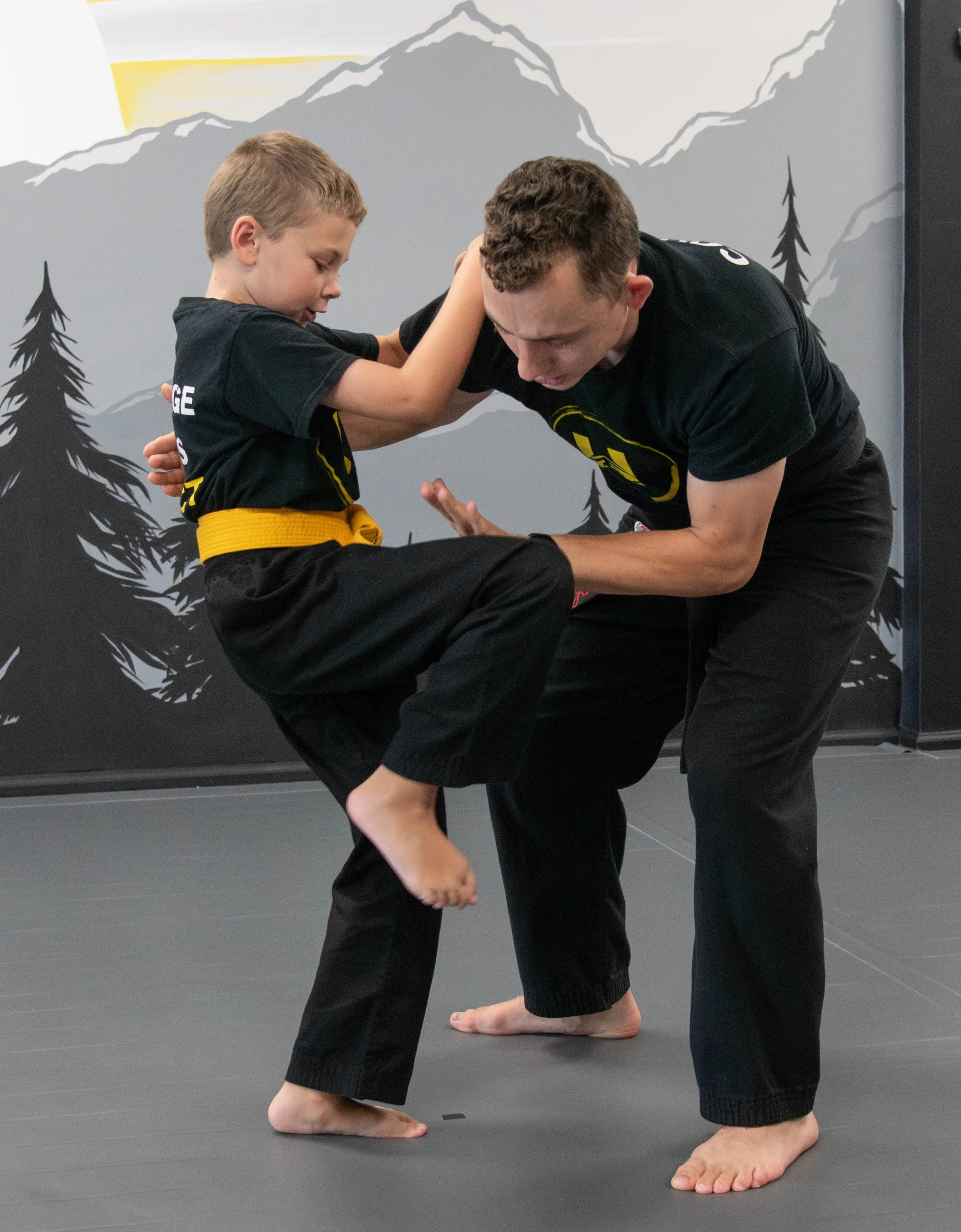 A young boy and an instructor practicing Brazilian Jiu-Jitsu on a mat with a mountain and trees mural in the background.