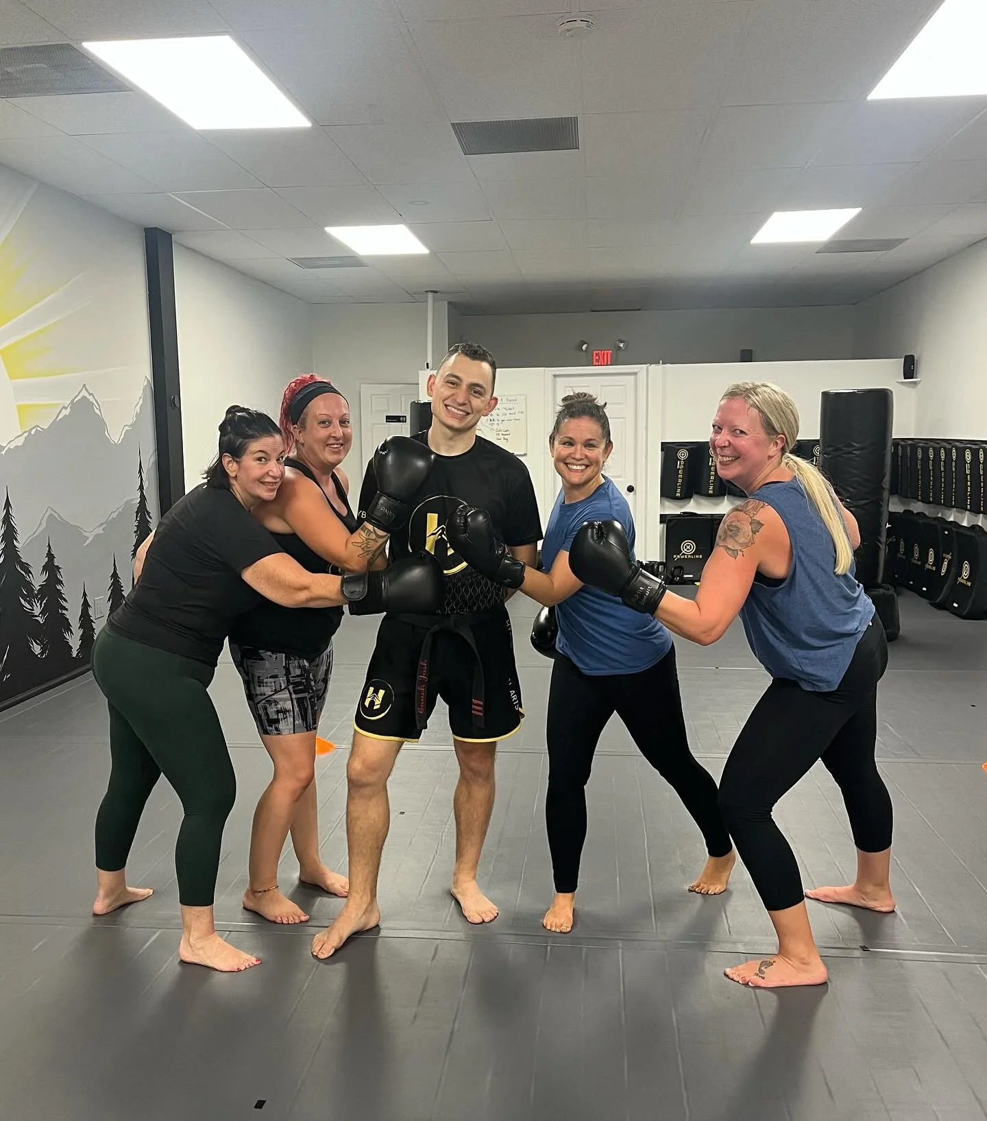 Group of five women and one man standing in a martial arts gym, wearing boxing gloves, smiling, and posing in a circle with their hands connected.