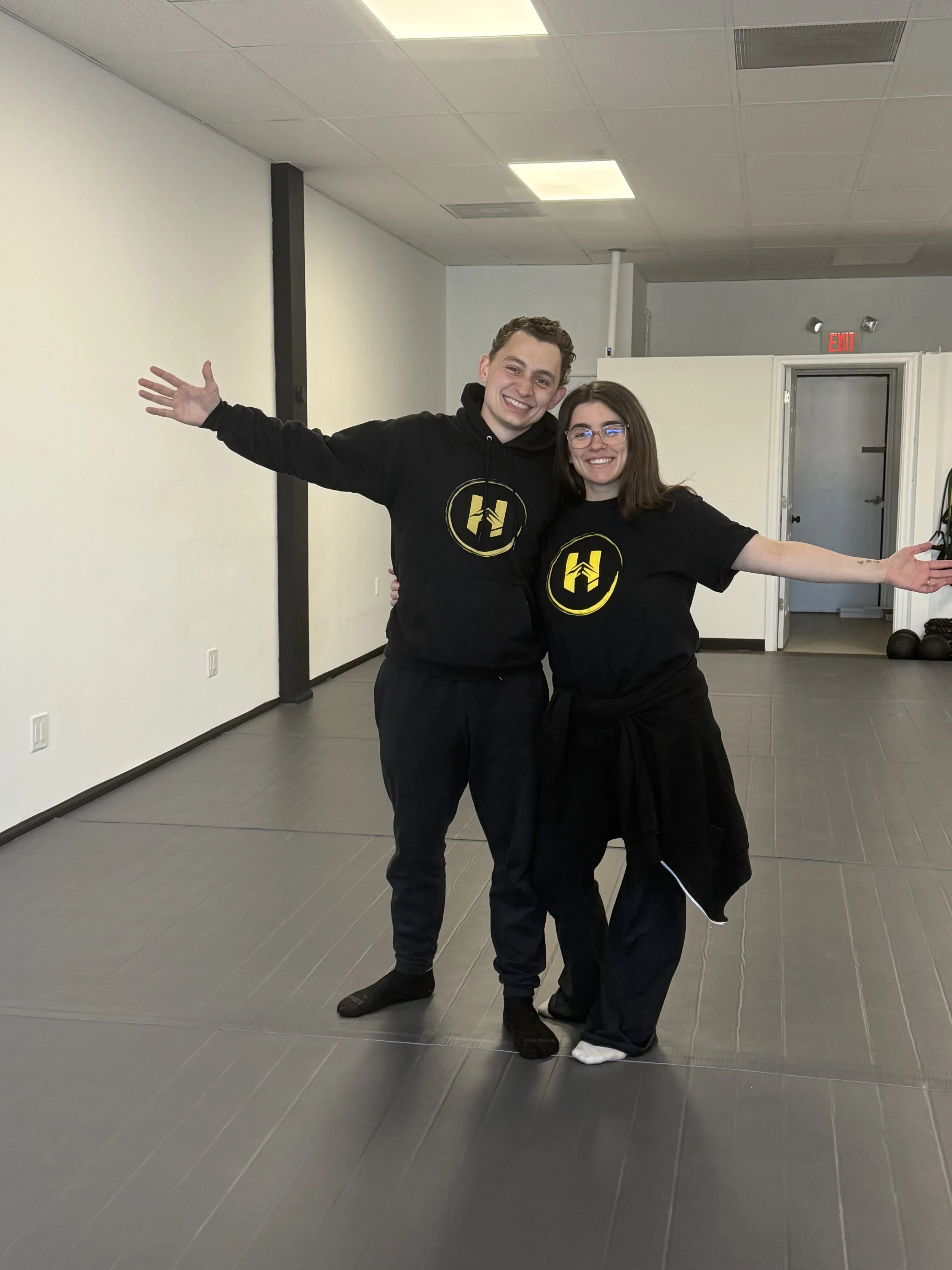 Two smiling people in black T-shirts with yellow logos, standing in a dance studio or gym with gray tile floors, white walls, and a drop ceiling, posing with arms outstretched.