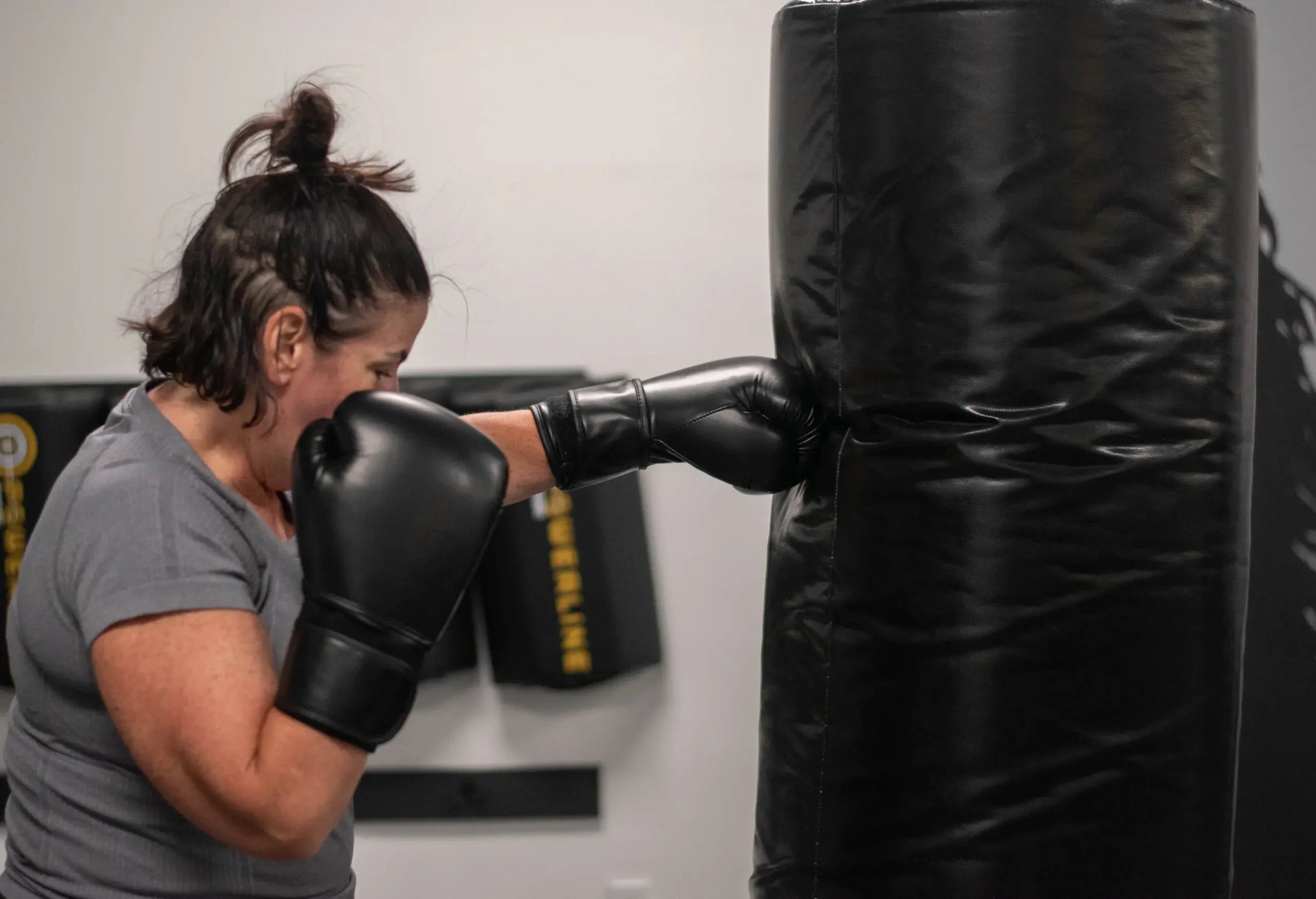 A woman in gray shirt wearing black boxing gloves practicing a punch on a black punching bag in a gym.