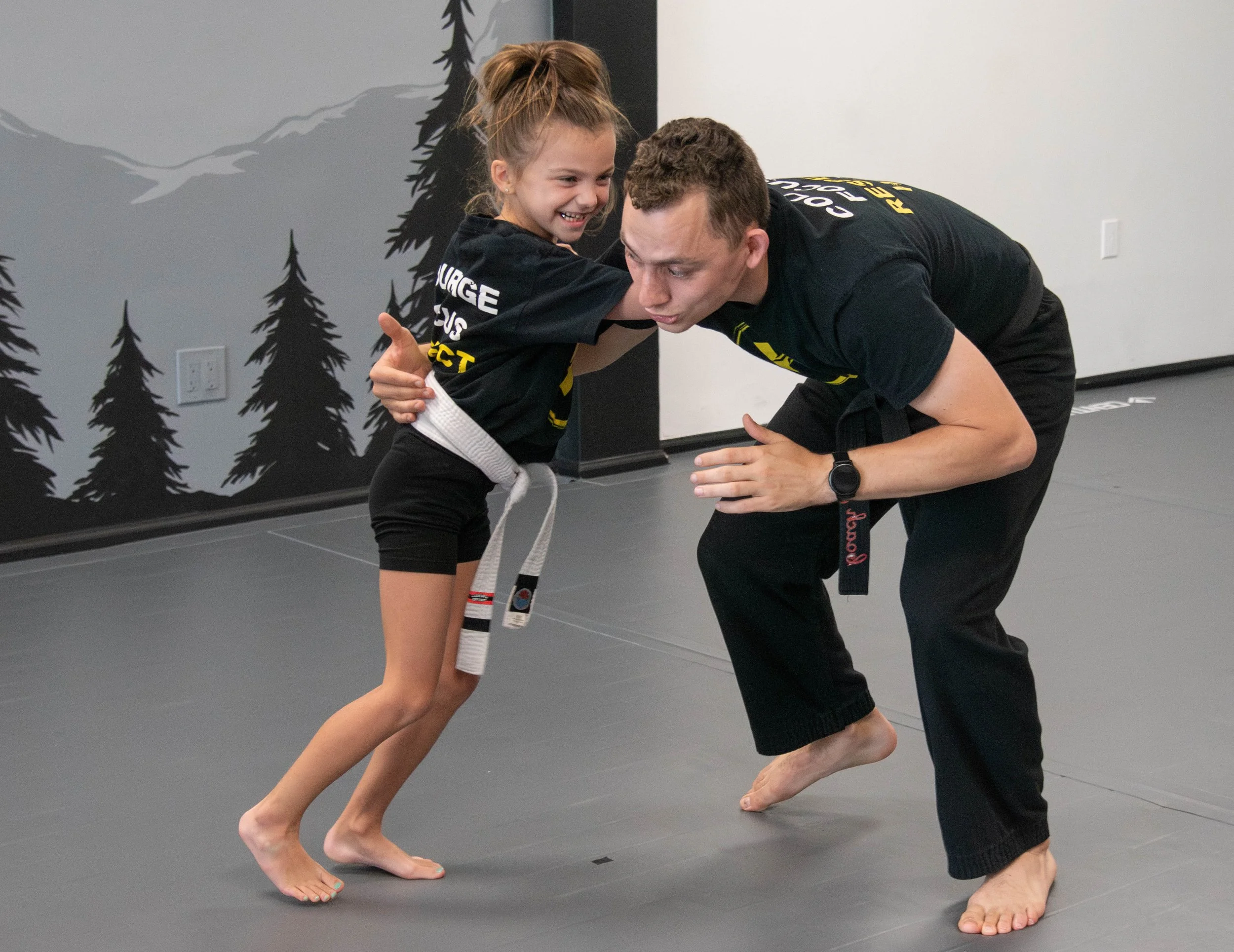Young girl in a martial arts uniform practicing Brazilian Jiu-Jitsu with her instructor in a dojo with a forest mural on the wall.