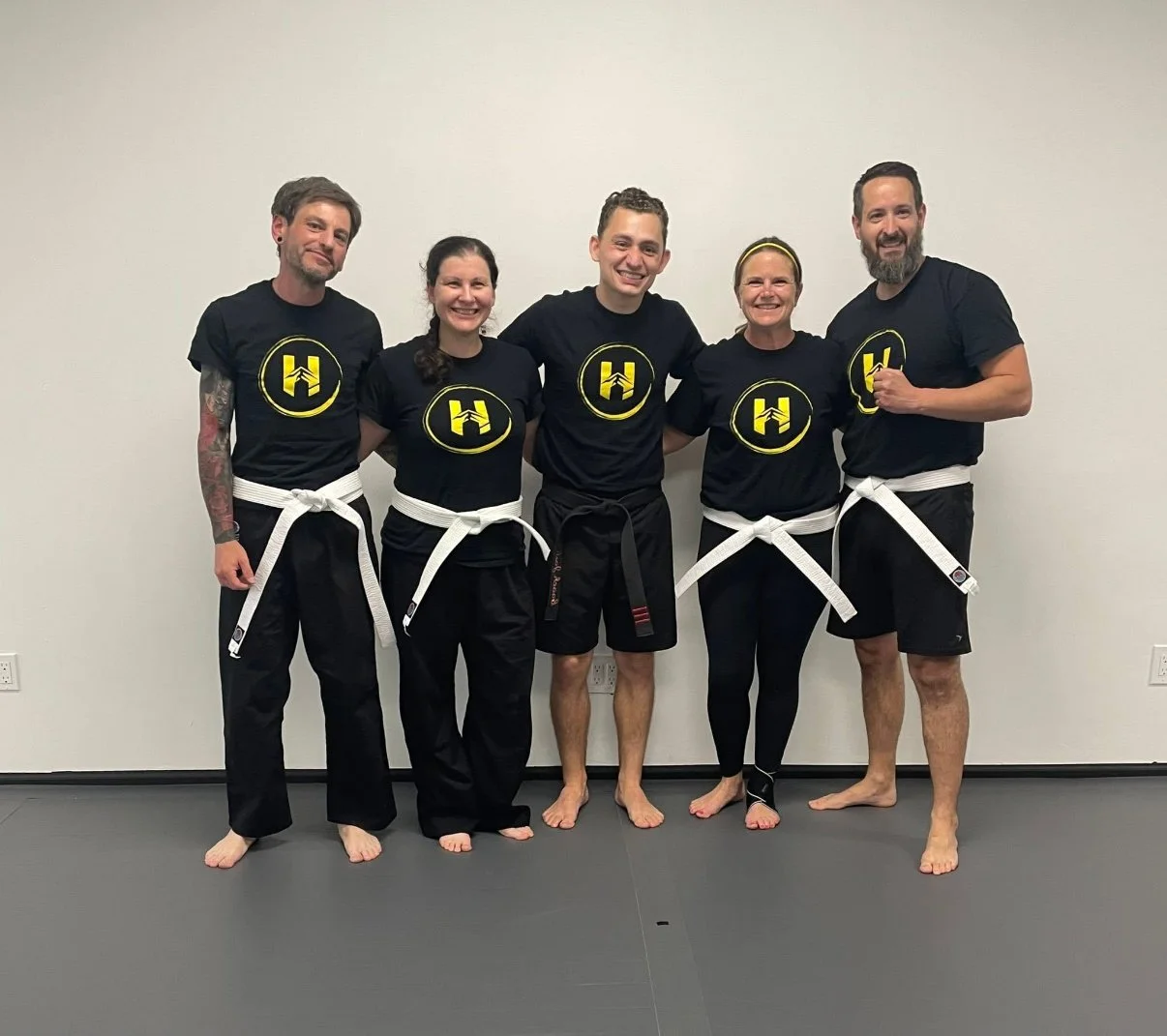 Group of five people in martial arts uniforms with white belts, standing on a gray mat against a plain white wall, smiling for the camera.