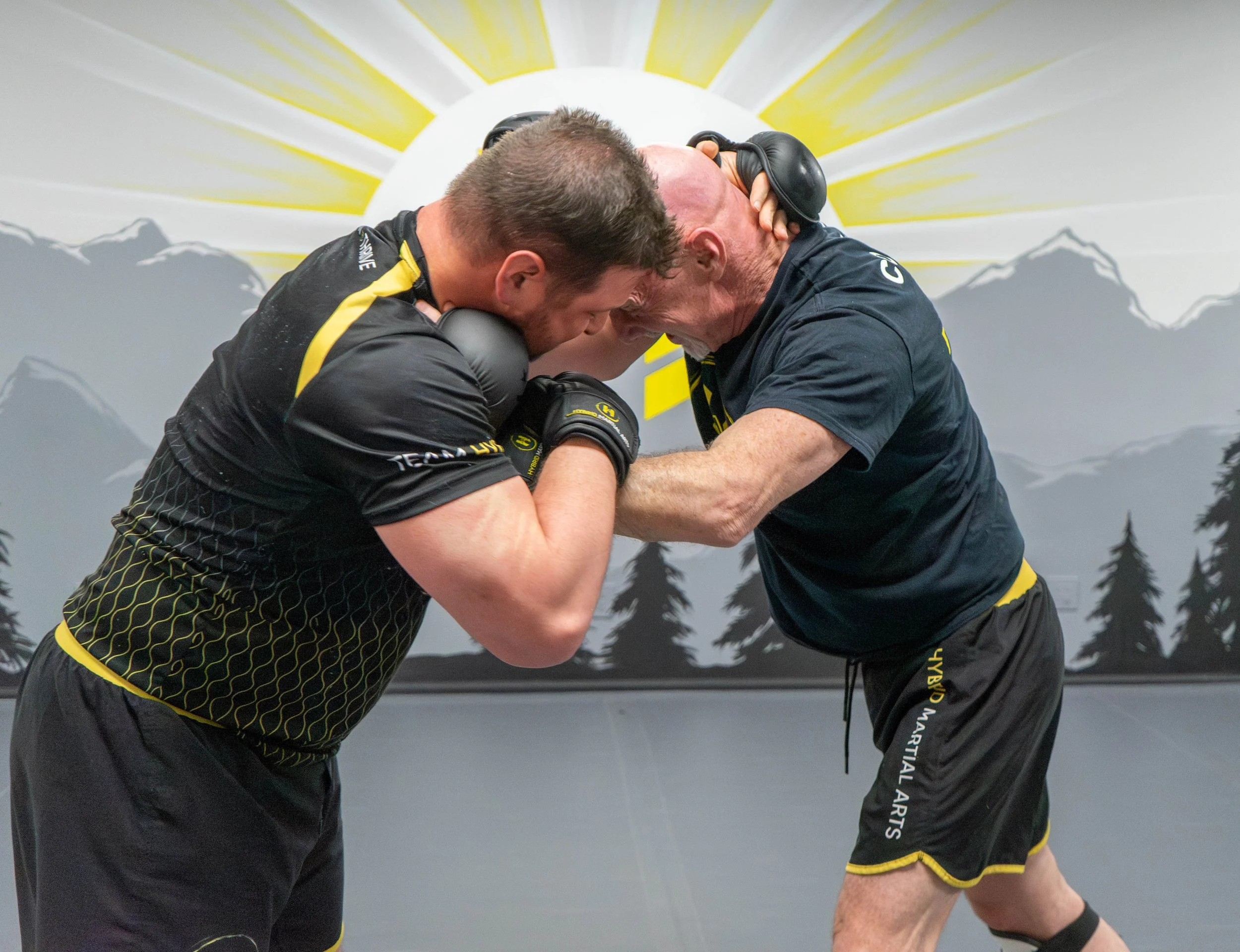 Two men practicing martial arts in a gym with mountain and forest mural background, wearing black shorts and gloves, engaging in a grappling technique.