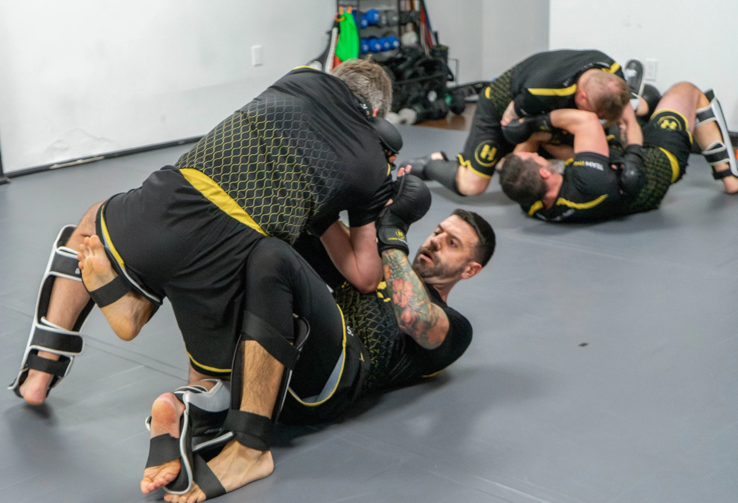 Two men grappling in a Brazilian Jiu-Jitsu training session, wearing black rash guards with yellow accents, on a gray mat in a gym.