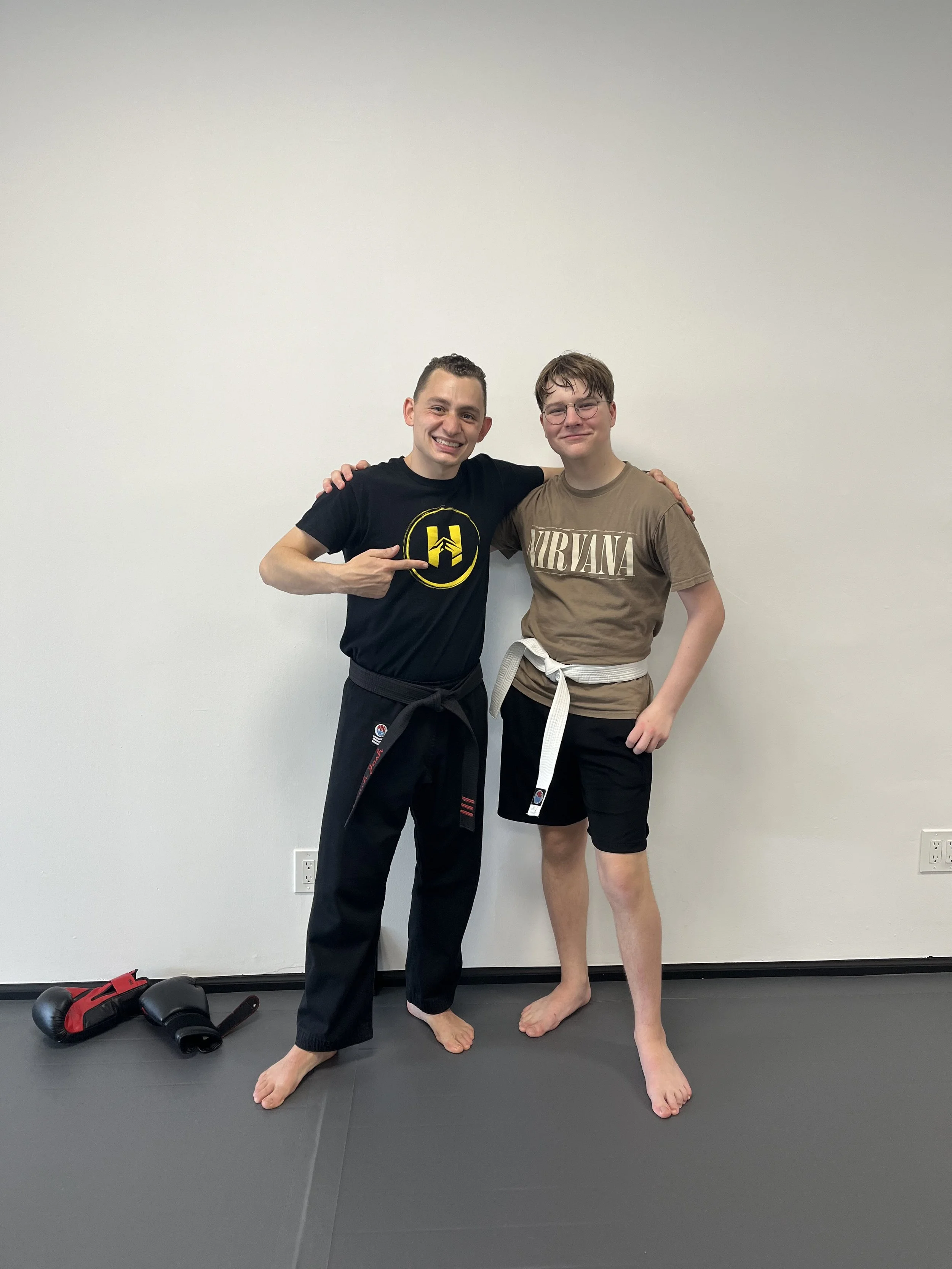 Two young men in martial arts uniforms standing together on a black mat against a plain white wall, smiling at the camera, with boxing gloves on the floor nearby.