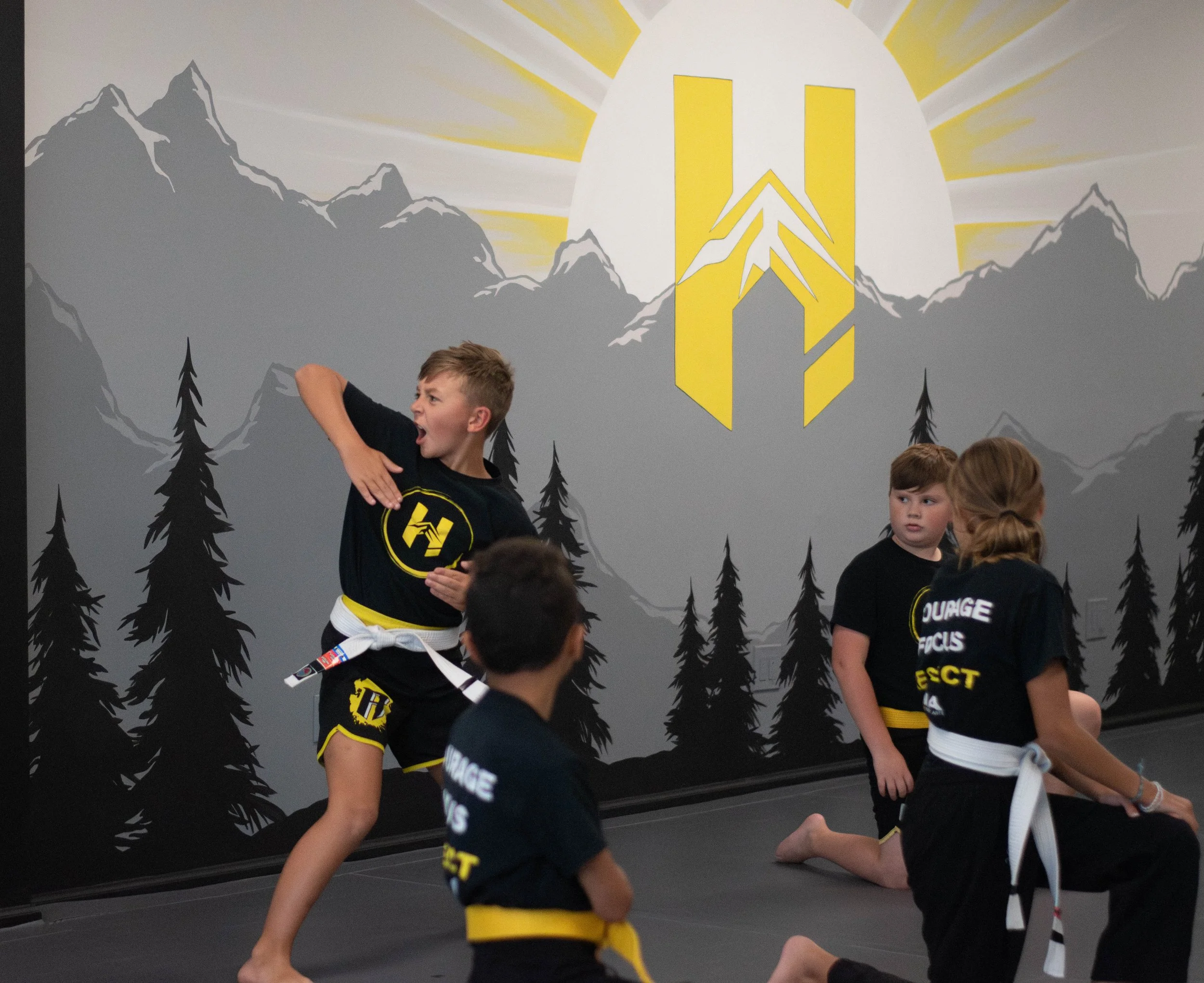 Children practicing martial arts in black uniforms with yellow details in a training room with gray mountain and pine tree mural behind them.
