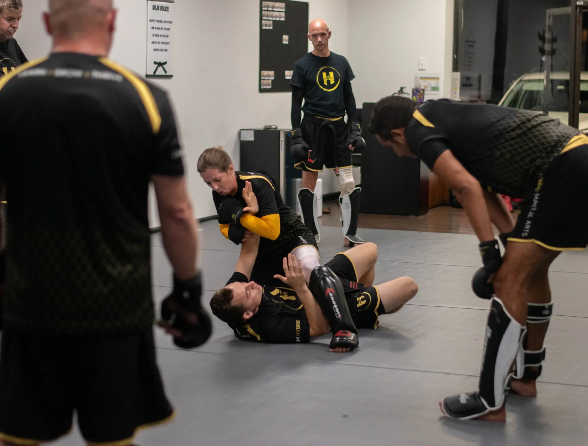 Martial arts training session with four students in black and yellow uniforms and a coach, practicing on gray mats in a gym.