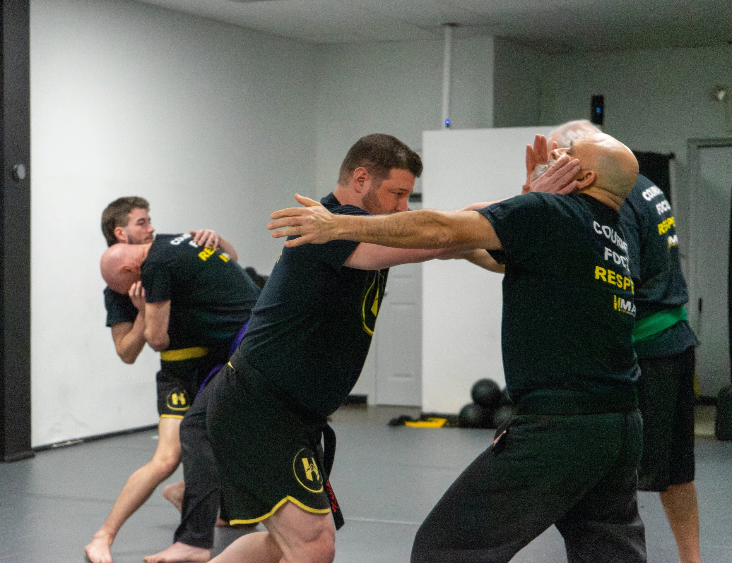 Two martial artists practice self-defense techniques on each other in a gym, wearing black T-shirts and shorts, with other participants training in the background.