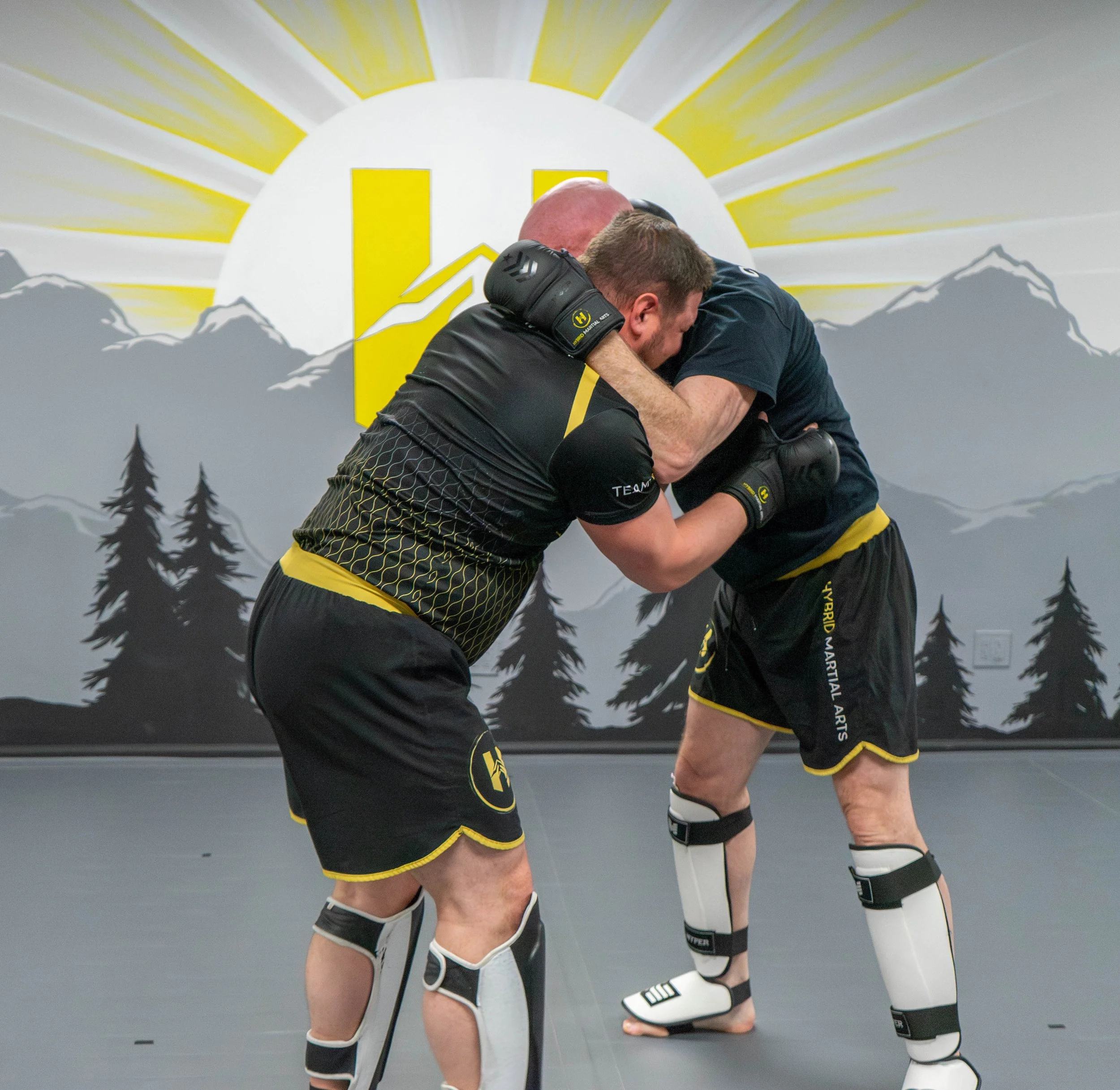 Two men in martial arts uniforms are engaged in a friendly hug in a gym, with a backdrop featuring a stylized sunrise, mountains, and trees.