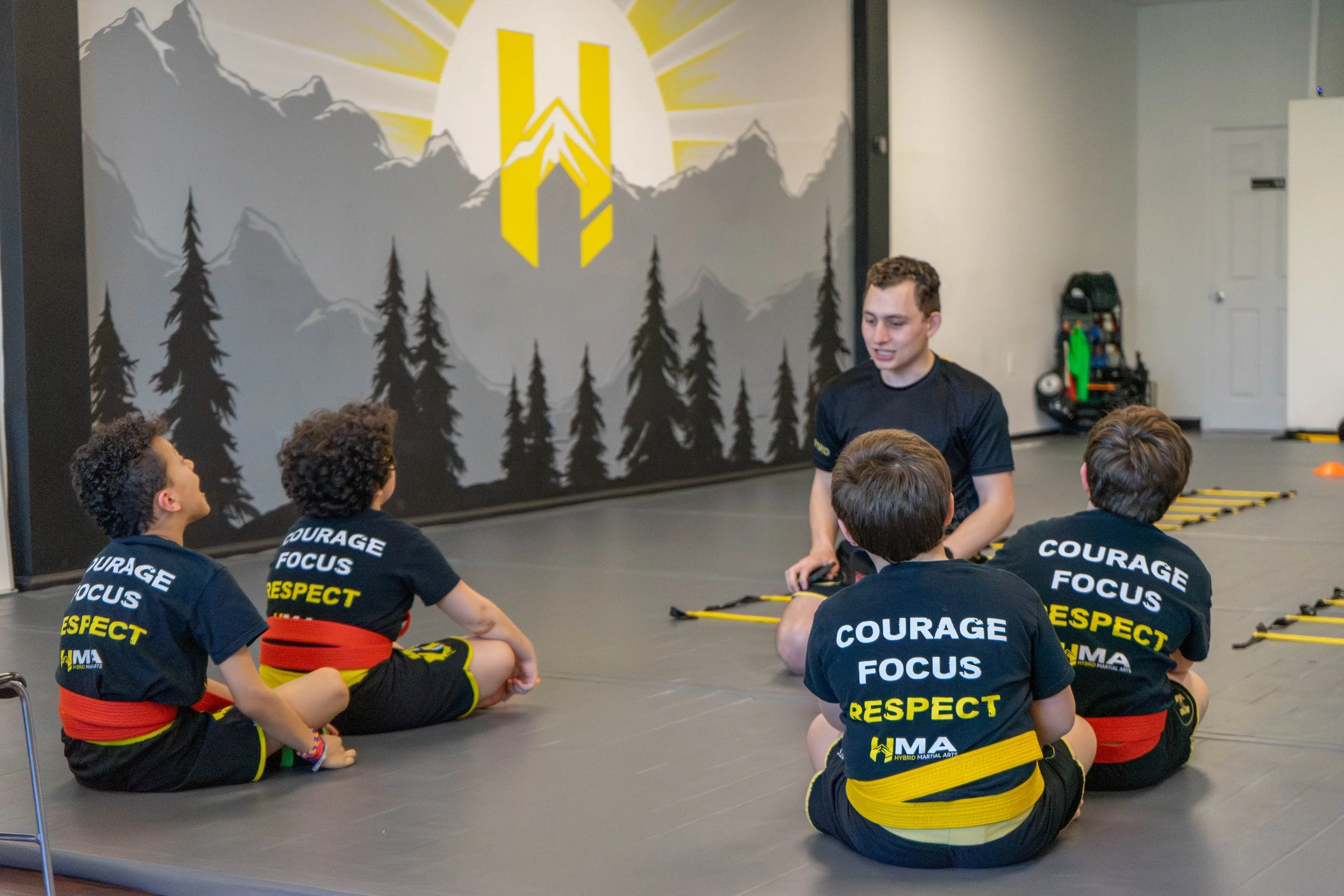 Young martial arts students sitting on the floor during a class, wearing black uniforms with words like 'COURAGE', 'FOCUS', 'RESPECT' printed on the back, in a martial arts studio with a mountain-themed mural on the wall.