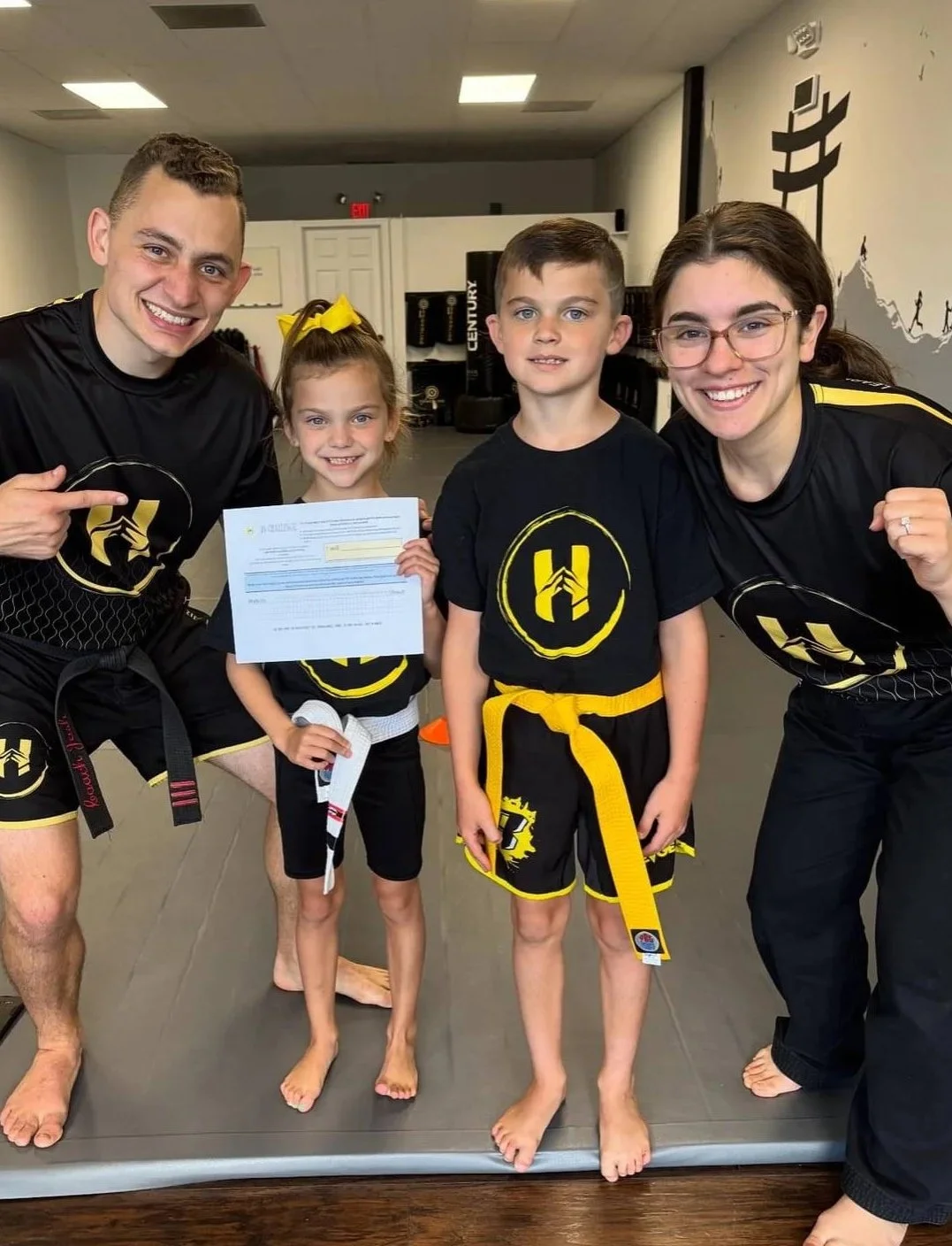 A group of four people, two children and two adults, at a martial arts dojo. The children are holding a certificate, and all are wearing black martial arts uniforms with yellow belts and a logo. The adults are pointing or flexing, and they are all standing on a martial arts mat.