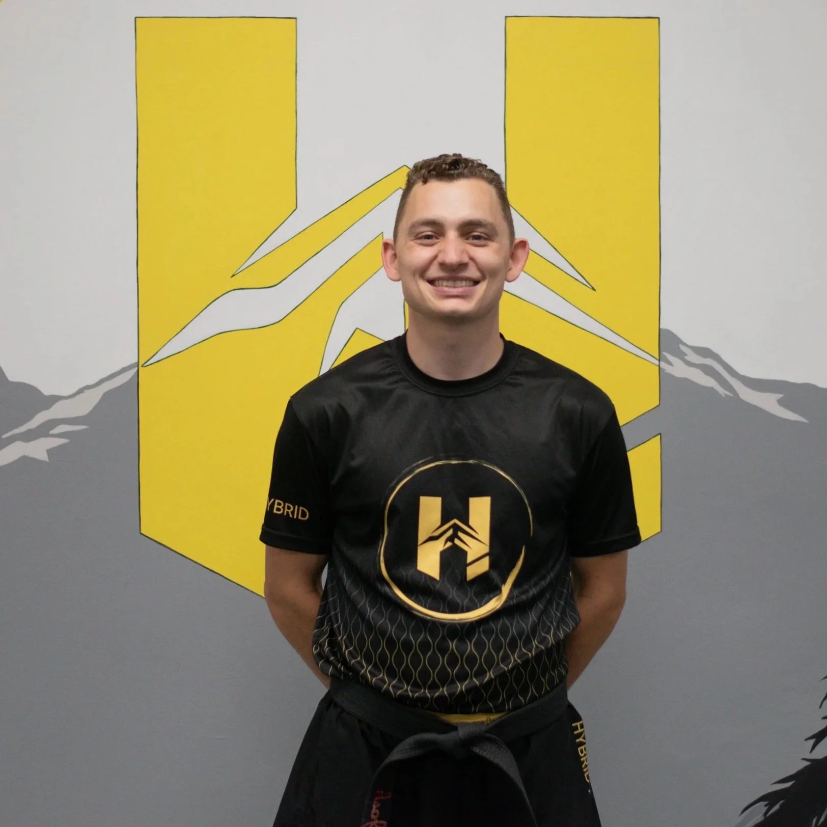A young man in a black martial arts uniform standing in front of a mountain-themed yellow and gray wall, smiling at the camera.