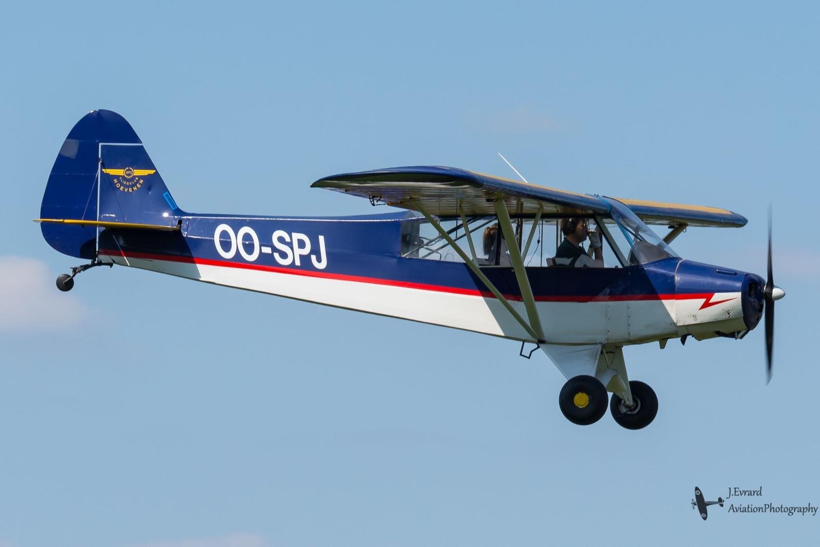 Een klein blauw en wit vliegtuig in de lucht met opdruk OO-SPJ, met twee mensen in de cockpit. Het is een propeller vliegtuig met een open cabine en er is een blauwe lucht op de achtergrond.
