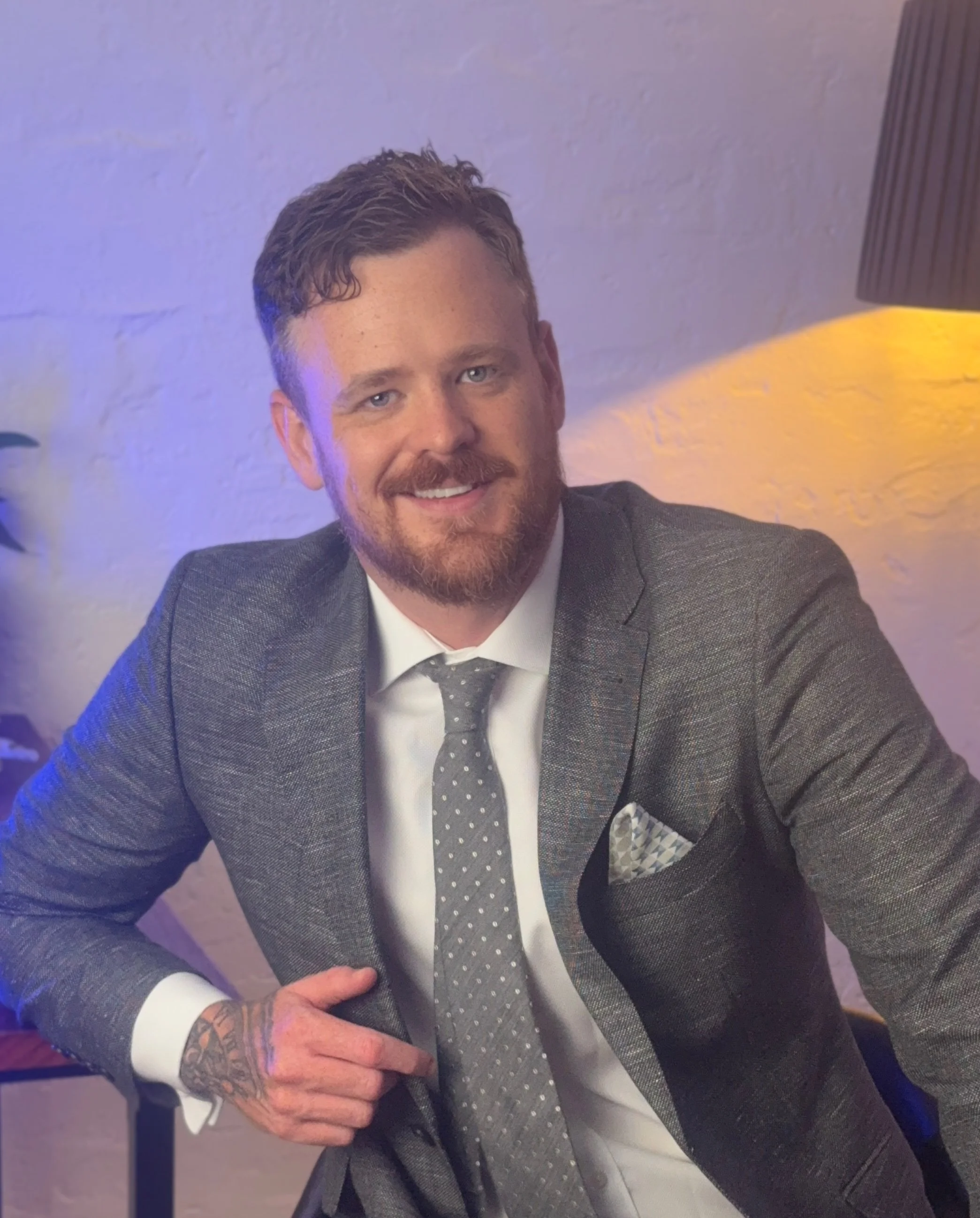 A man with red hair and a beard, dressed in a gray blazer, white shirt, and patterned tie, sitting with one hand resting on his knee. The background features a textured white wall and a lamp casting warm yellow light.