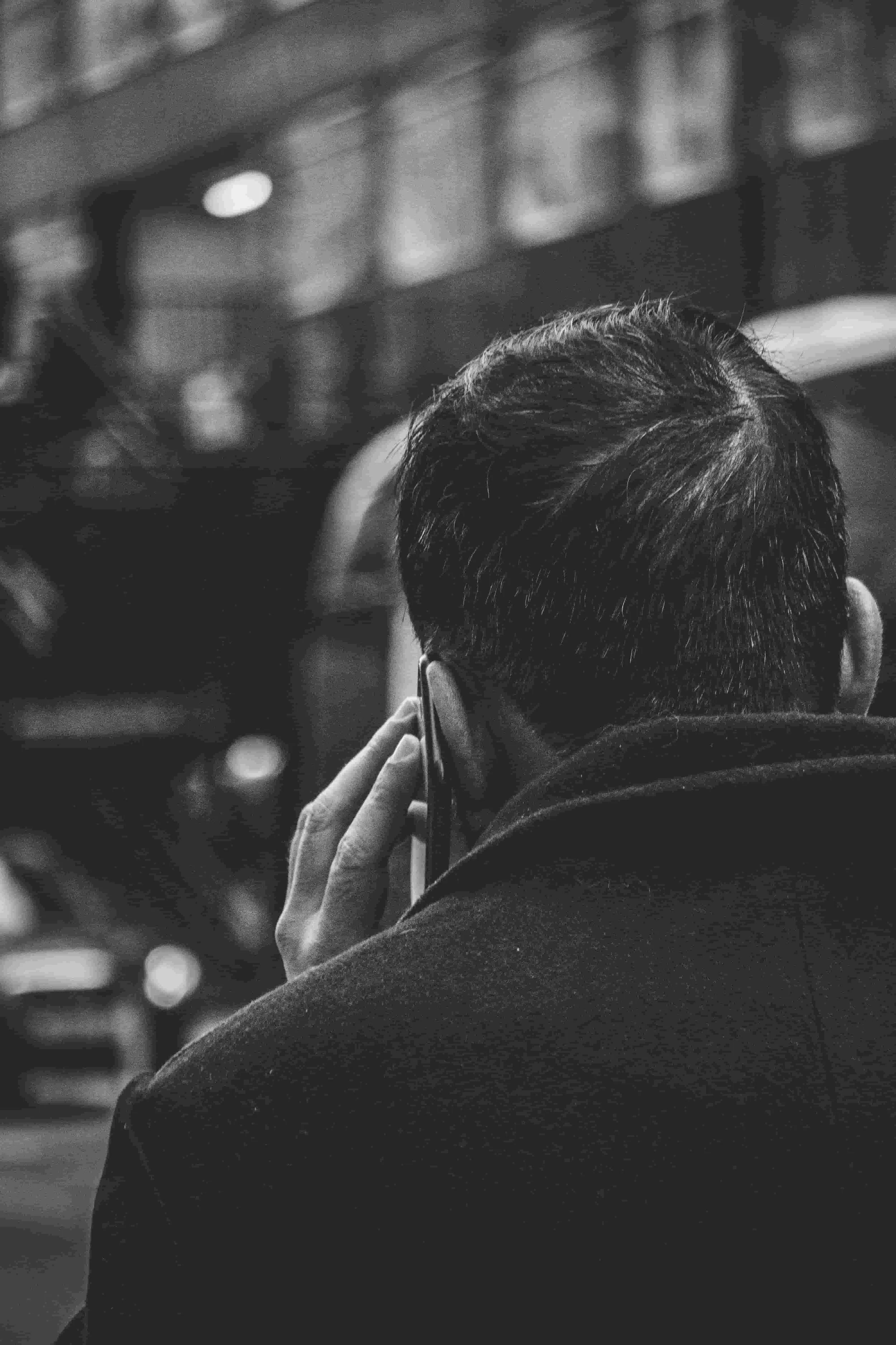 A person with dark hair and glasses is holding a phone to their ear, viewed from behind. The scene is in black and white with a blurred background.