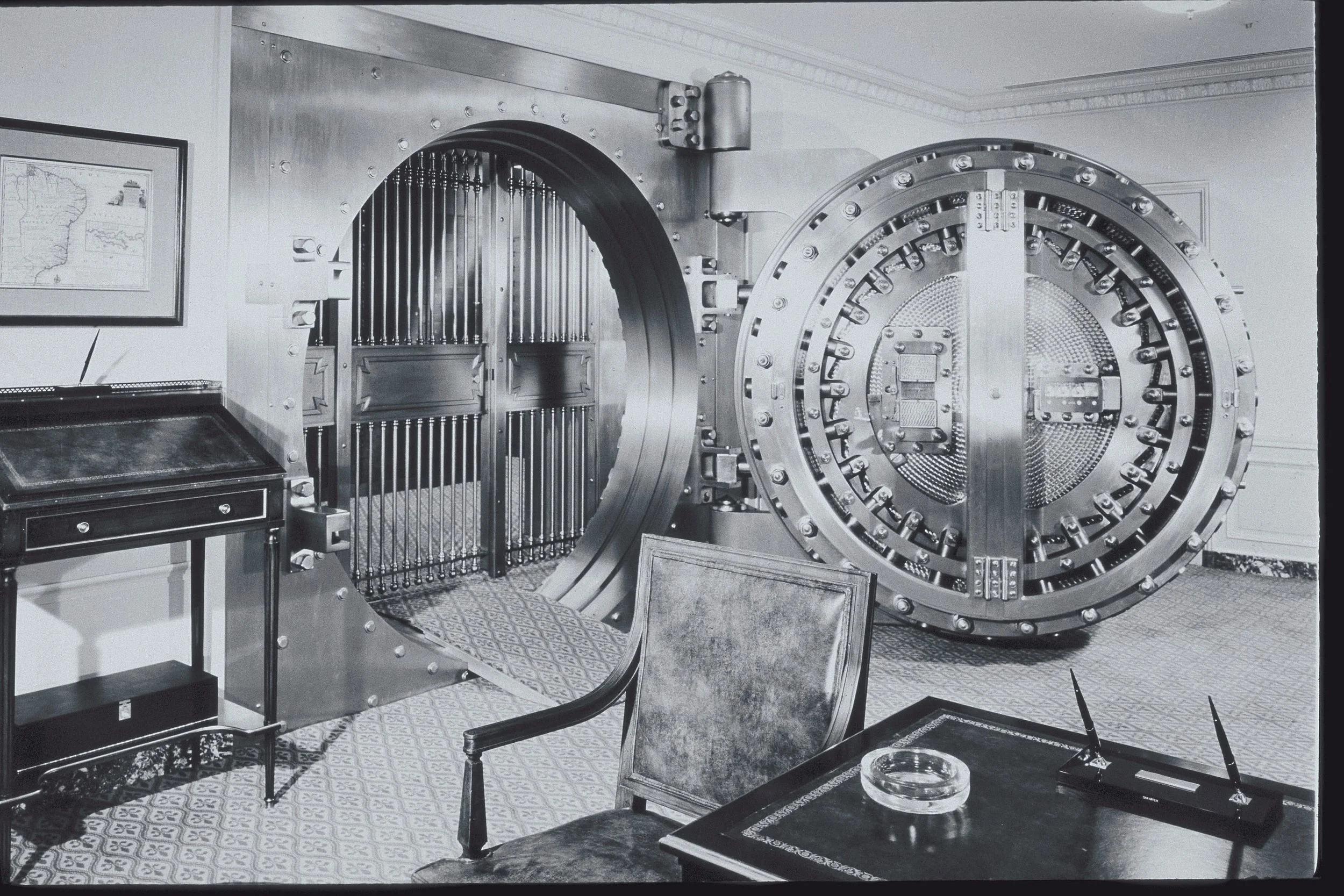 Black and white photograph of a large, open bank vault door in a room with patterned carpet and decorative ceiling. The vault door is round with multiple bolts and a central locking mechanism. Nearby is a framed map on the wall and a small desk with a chair, an ashtray, and a telephone.