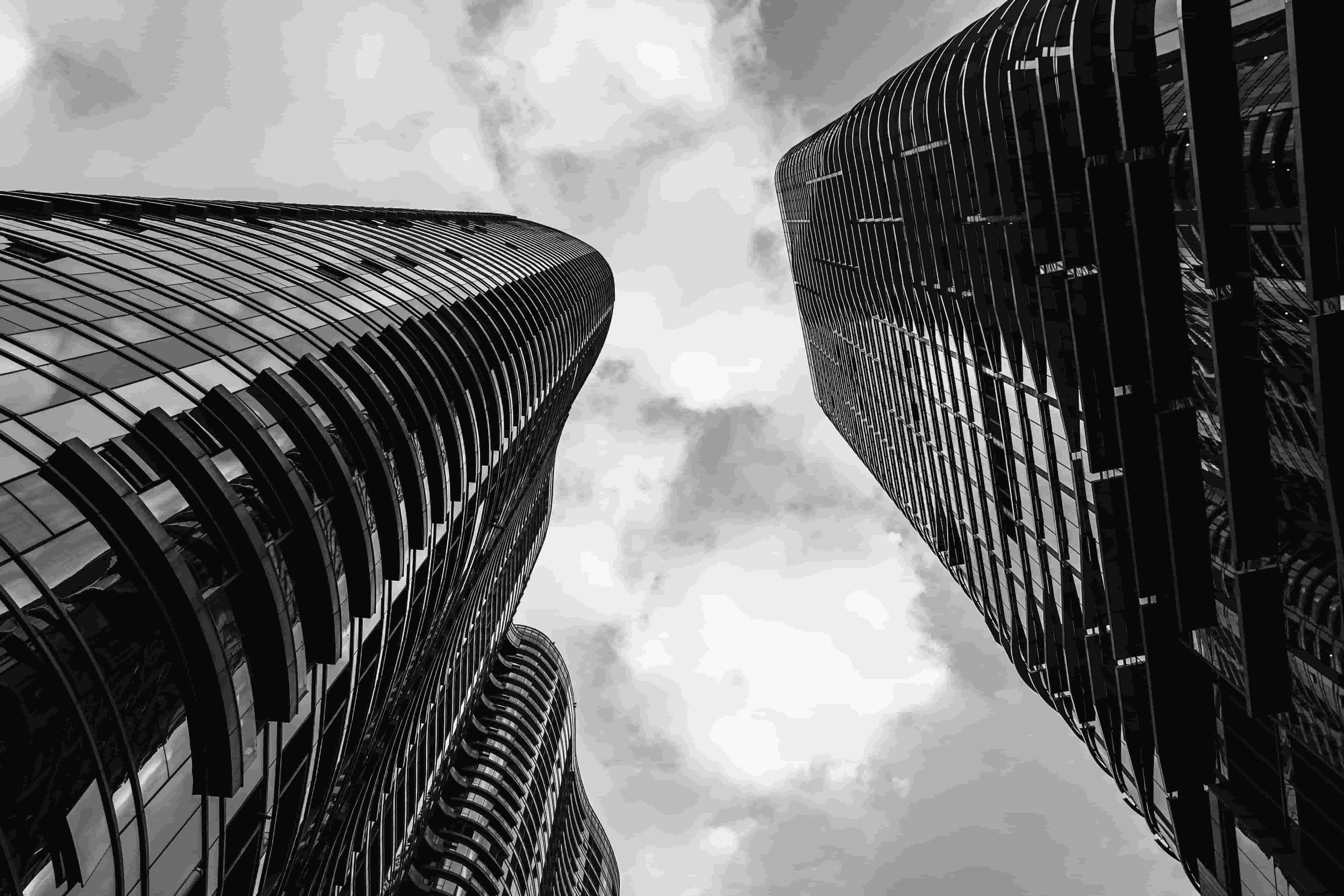 Two tall modern skyscrapers with curved glass facades reaching into a cloudy sky, seen from below.