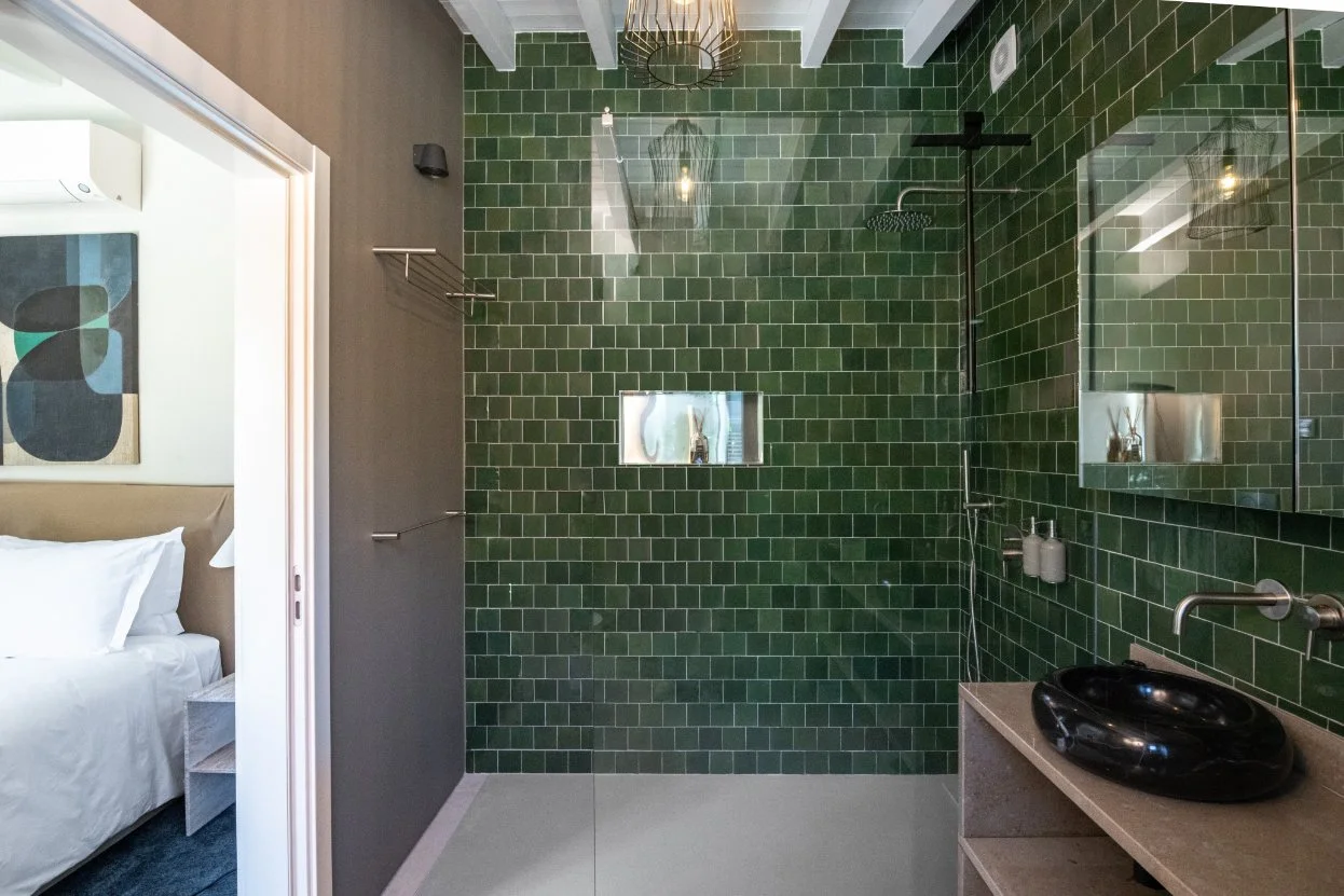 Modern bathroom with green subway tile walls, a glass shower enclosure, a black vessel sink on a beige countertop, and a mirror. Part of a bedroom with white bedding is visible through an open door.  Holiday rental home in Lagoa.