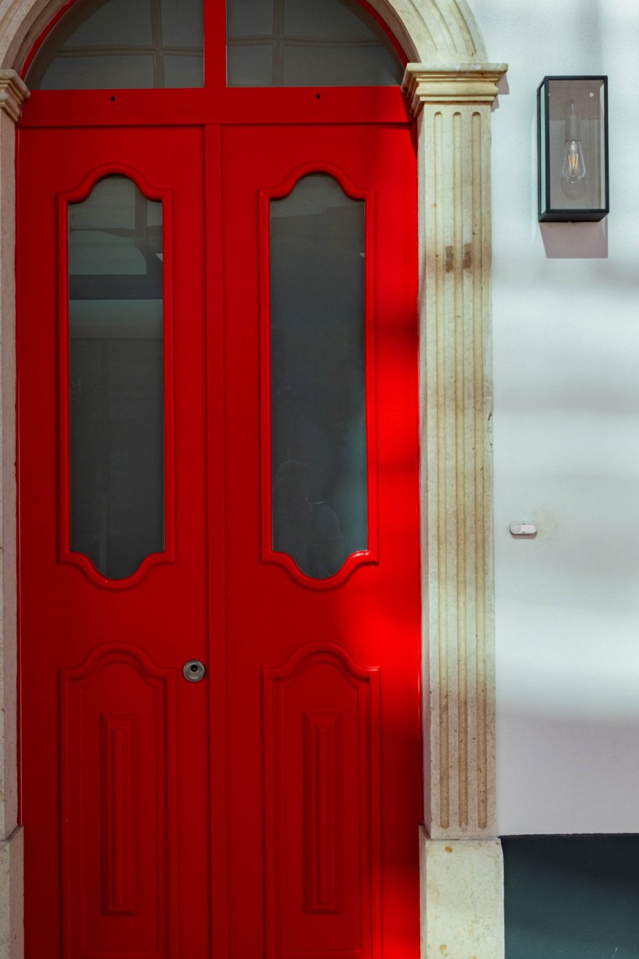 Red double doors with glass panels, framed by stone columns and a white wall with a modern wall-mounted light fixture.  Holiday rental home in Lagoa.