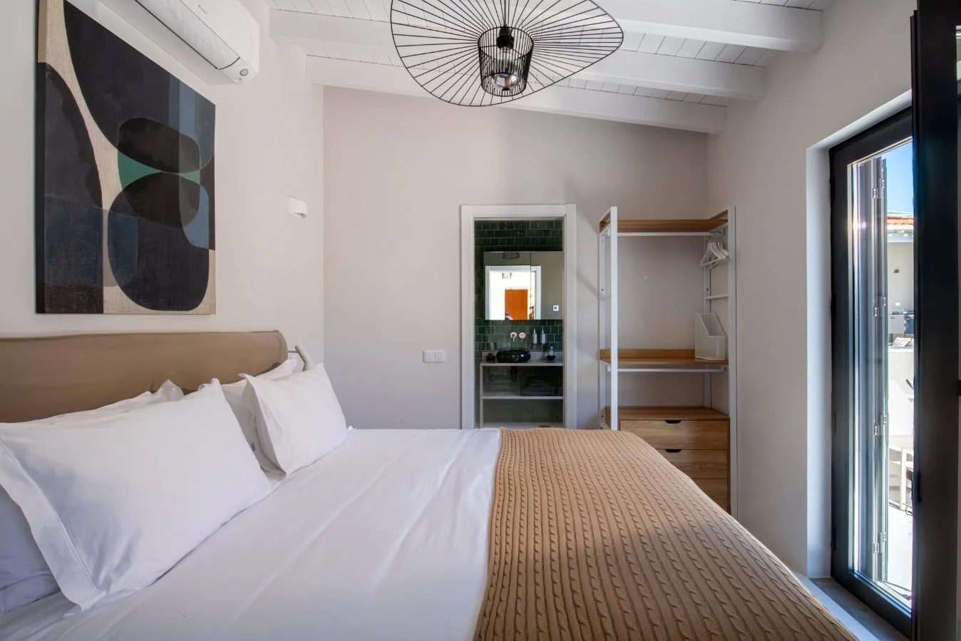 A bedroom with a bed that has white sheets and pillows, an artwork on the wall, and a sliding glass door leading outside. There's also a wooden open closet and a view into a bathroom with green tiles.  Holiday rental home in Lagoa.