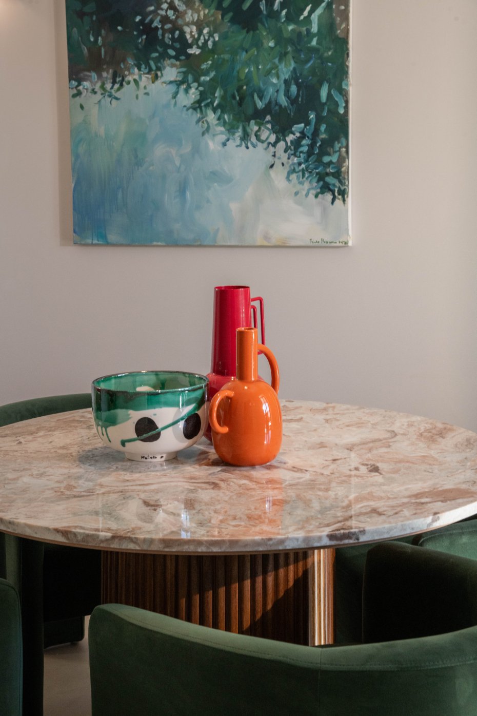 A round marble table with green velvet chairs around it. On the table are three ceramic vases in red, orange, and green, and a green and white patterned bowl. A large abstract painting with green and blue tones hangs on the wall behind the table.  Ho