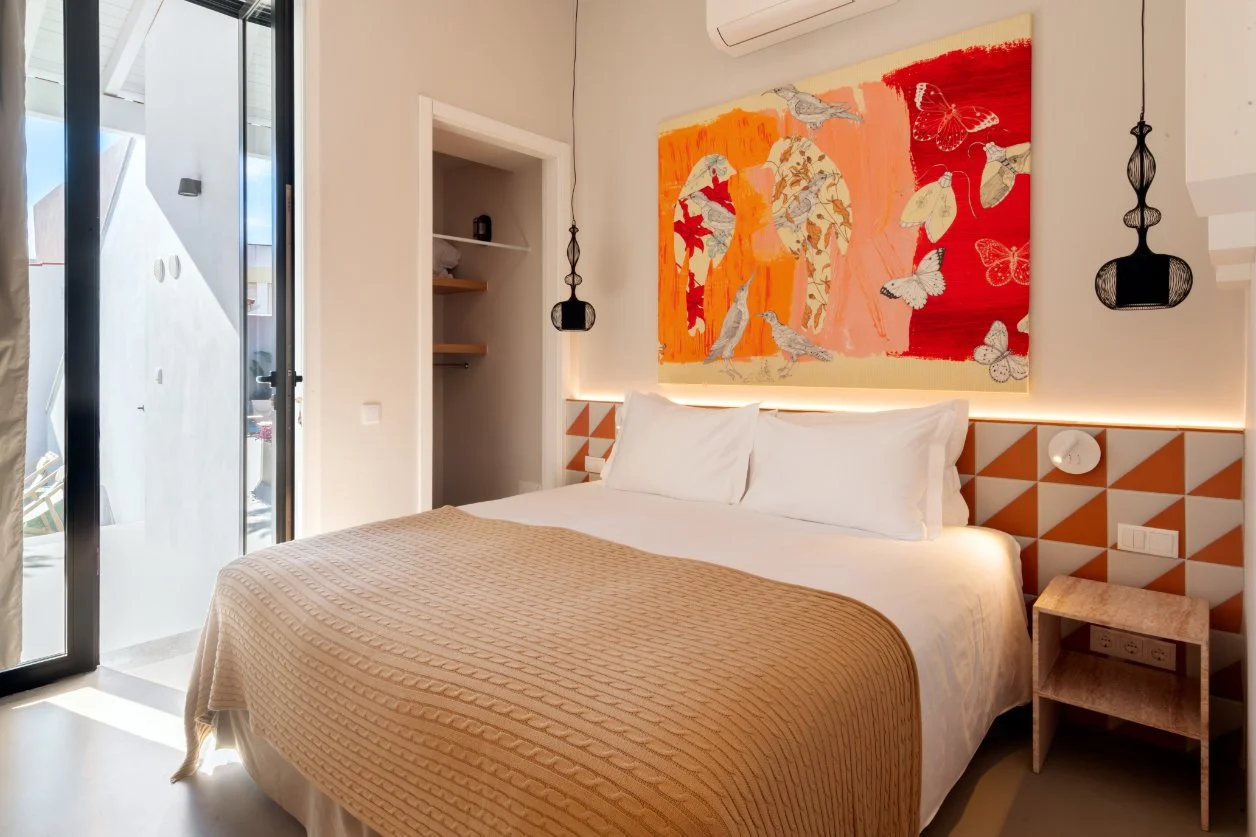 Bedroom with a large bed covered in white sheets and a beige bedspread, decorative orange and beige geometric headboard, colorful abstract art with birds and butterflies on the wall, small bedside table, black pendant lights.  Holiday rental home in 