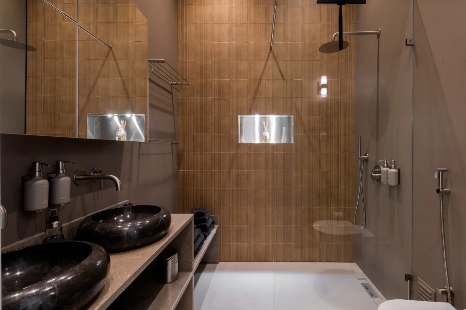 Modern bathroom with two black circular vessel sinks, tan countertop, wall-mounted faucets, large mirror, brown tiled walls, open shower area with rainfall showerhead, handheld shower, and built-in niche with toiletries.  Holiday rental home in Lagoa