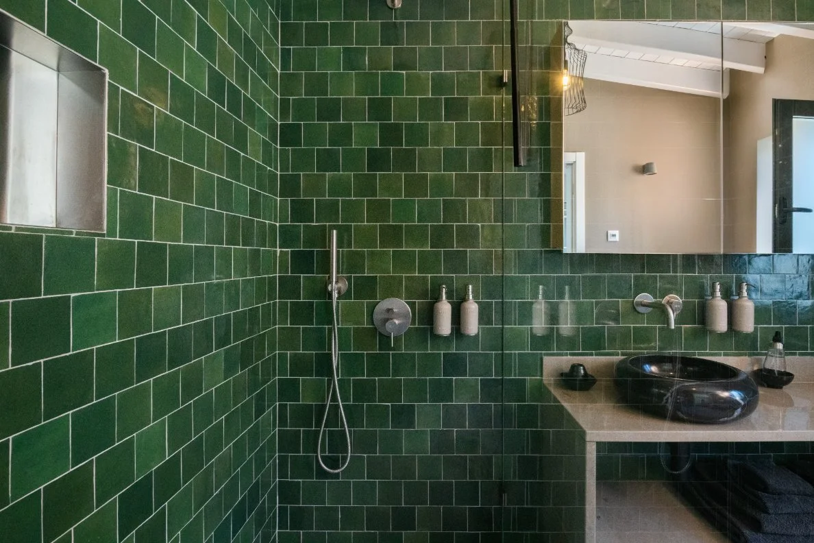 Bathroom with green tiled walls, a black sink, a mirror, and a showerhead with two bottles nearby.  Holiday rental home in Lagoa.