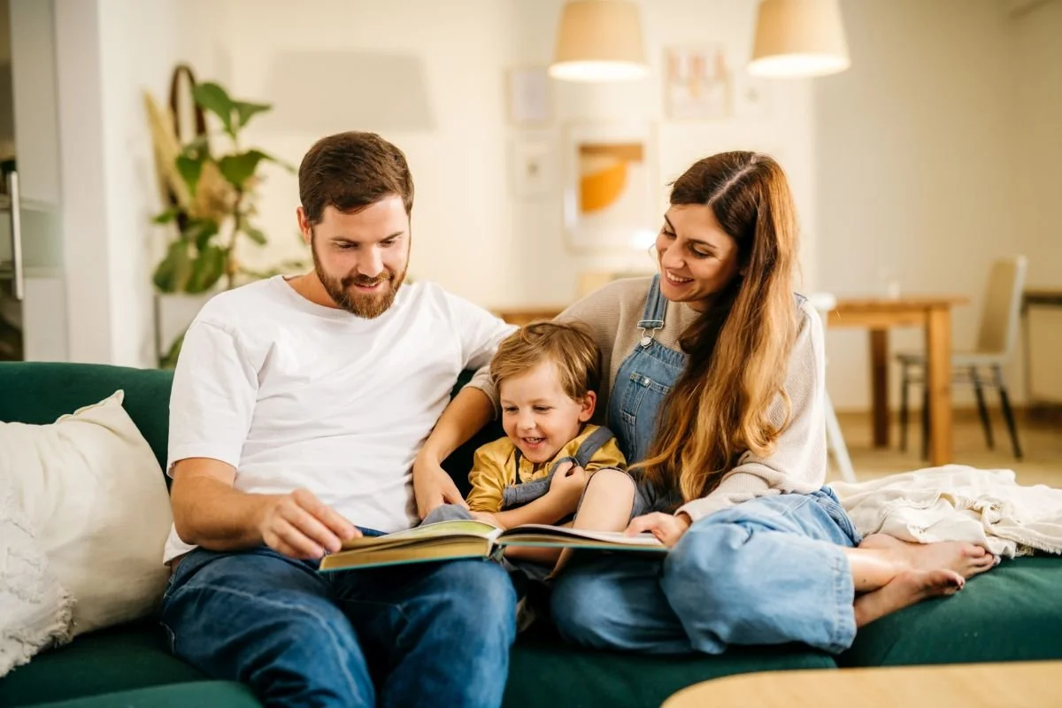 Eltern lesen ihrem kleinen Sohn auf dem Sofa ein Bilderbuch vor – Kind zu Hause in der Logopädie unterstützen