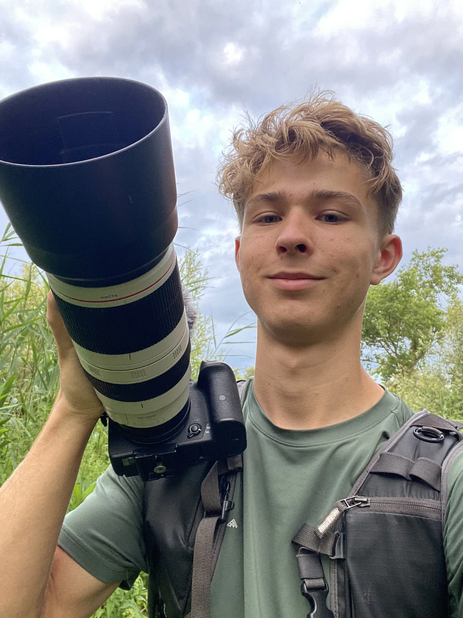 Young man with curly blond hair looking at camera, outdoors, holding a camera with a large lens, wearing a green shirt and a black backpack, green trees and a cloudy sky in the background.