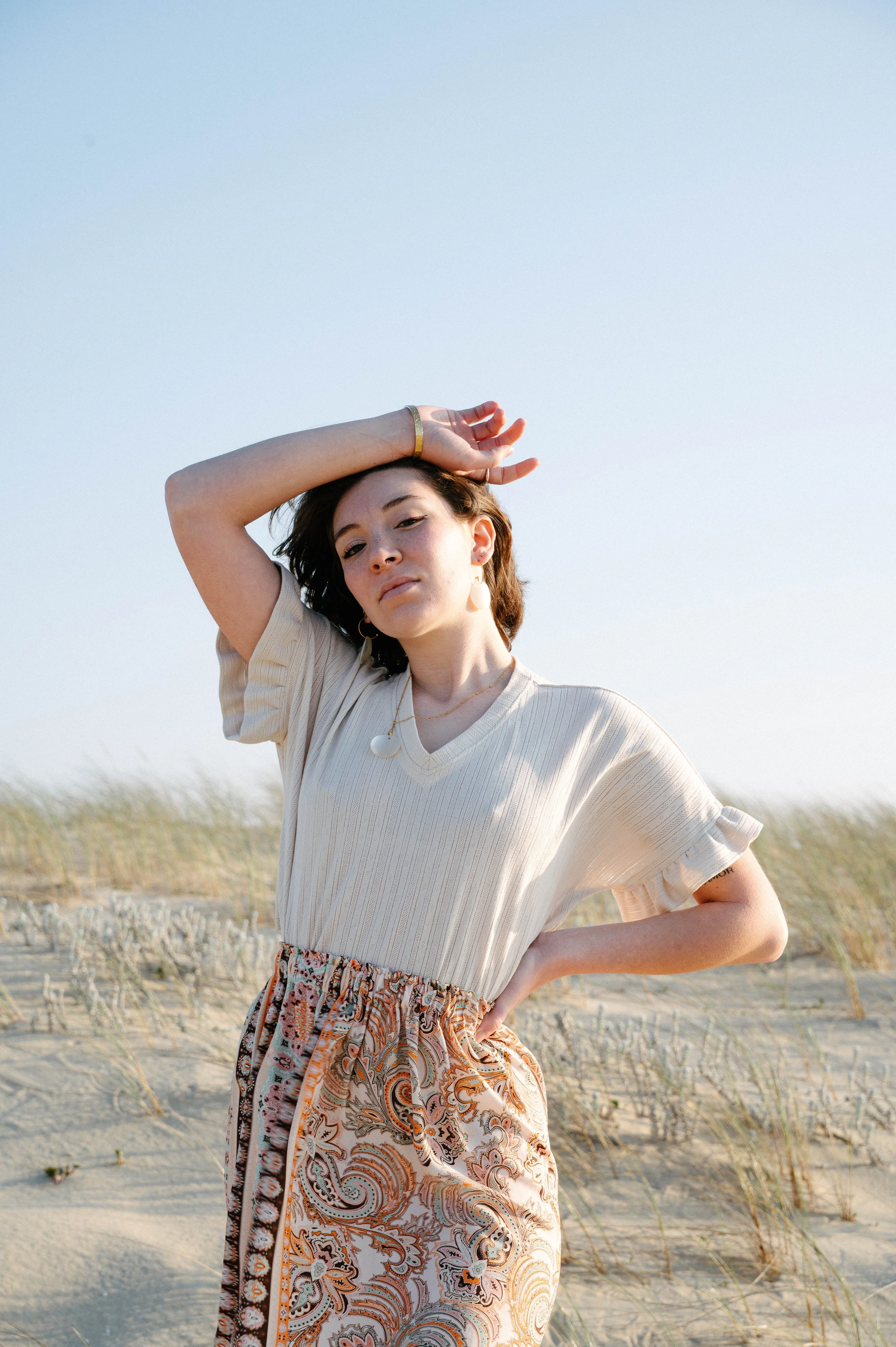 Femme en robe beige et jupe imprimée sur la plage, bras levé, paysage de dunes.