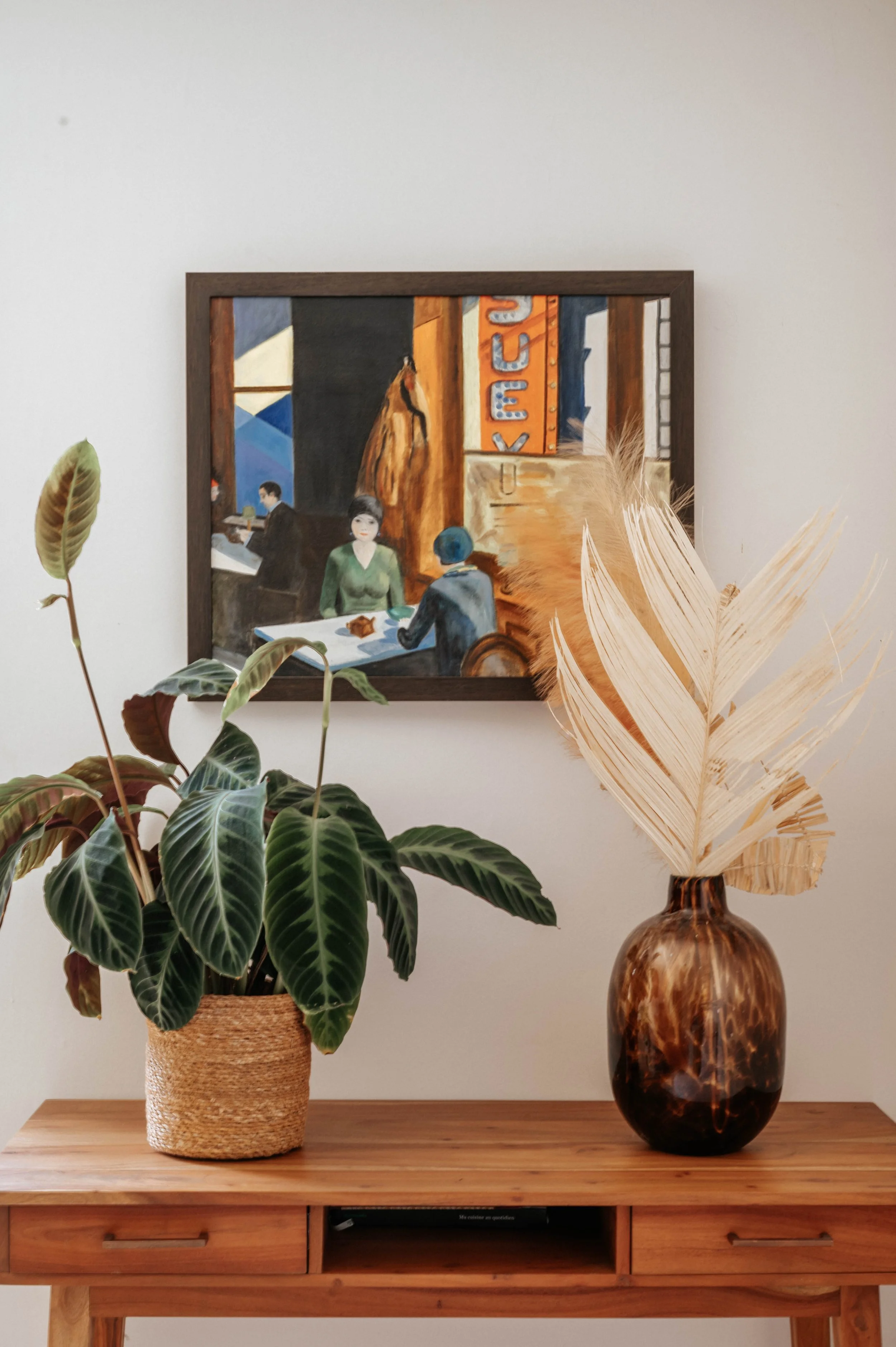 Table en bois avec plante en pot à gauche et vase avec feuilles séchées à droite. Tableau accroché au mur en arrière-plan.