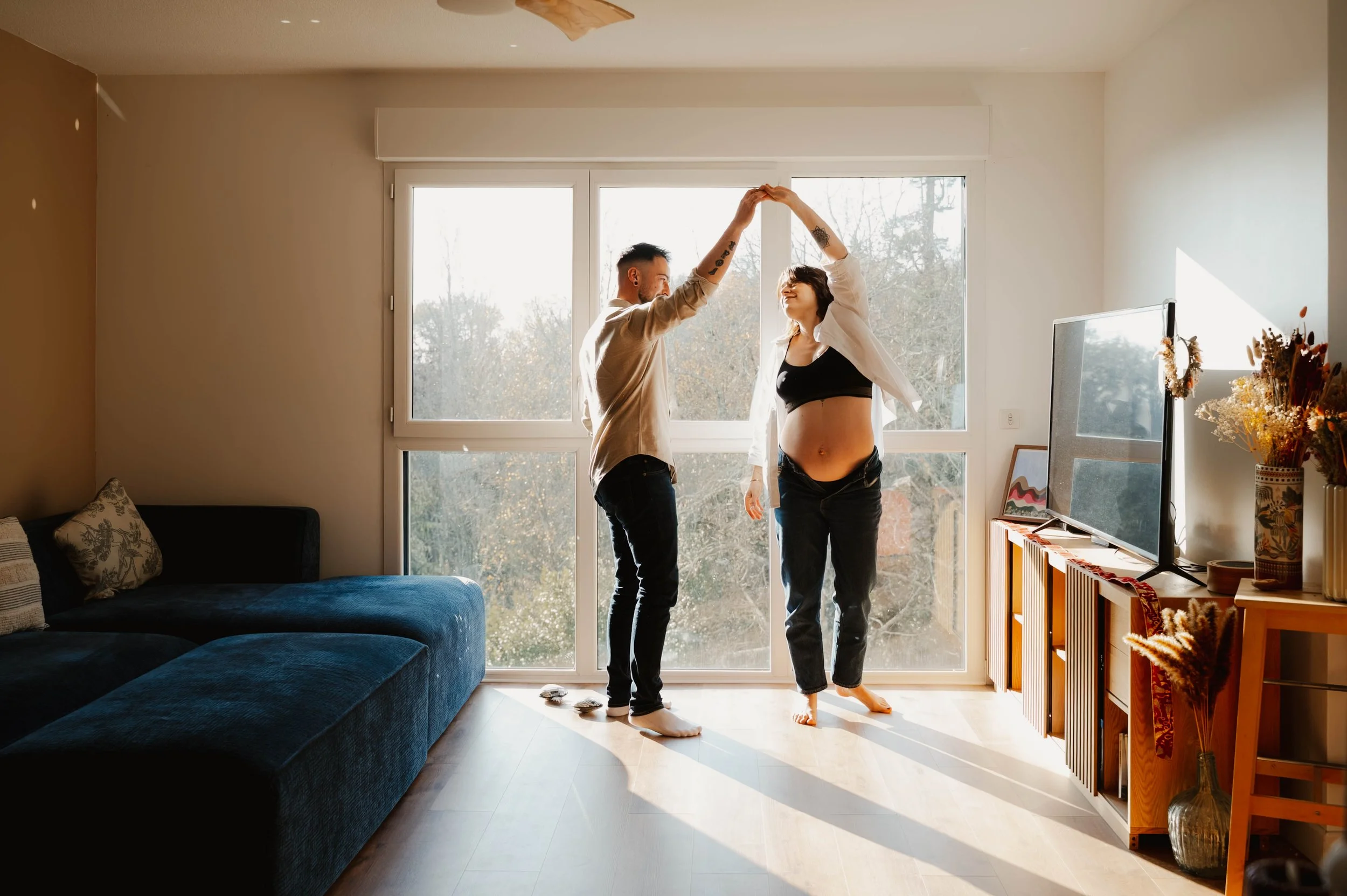 Un couple dans un salon, dansant ensemble, avec la femme qui a un ventre rond. La pièce est lumineuse avec de grandes fenêtres et des décorations florales sur un meuble.