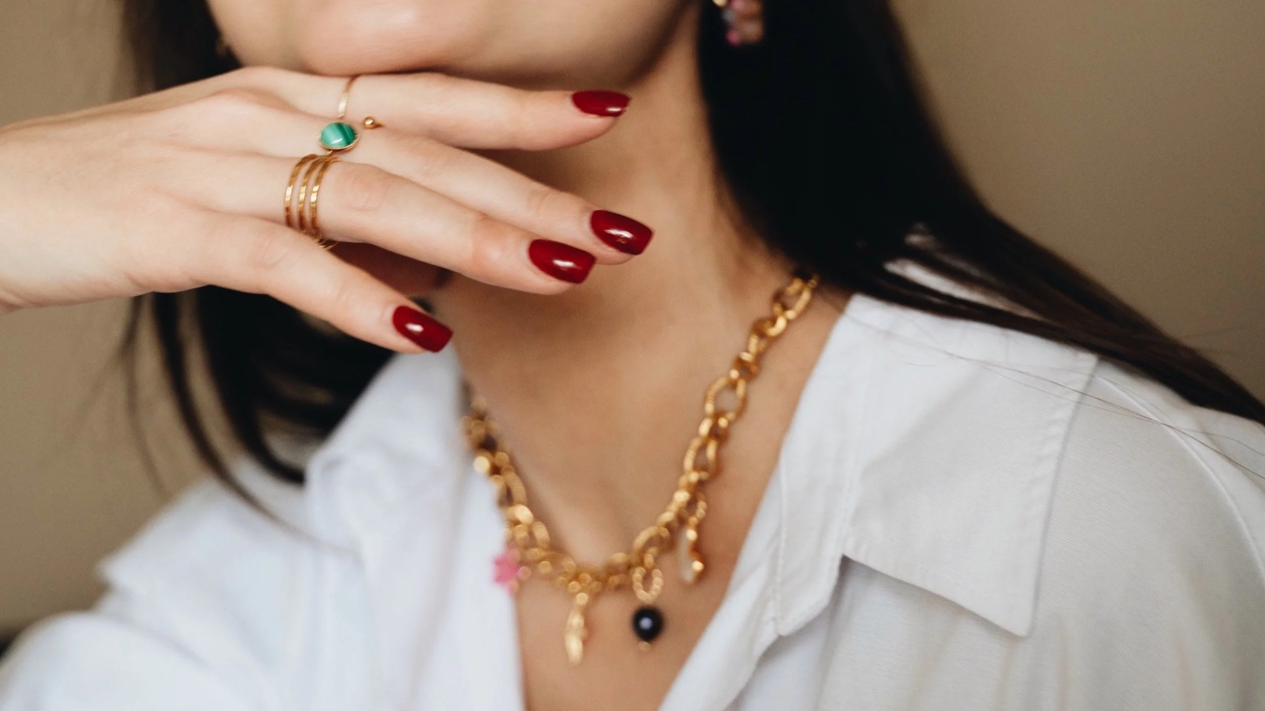 Femme portant un collier doré avec pendentifs, une chemise blanche et des bagues dorées, ongles vernis rouge.