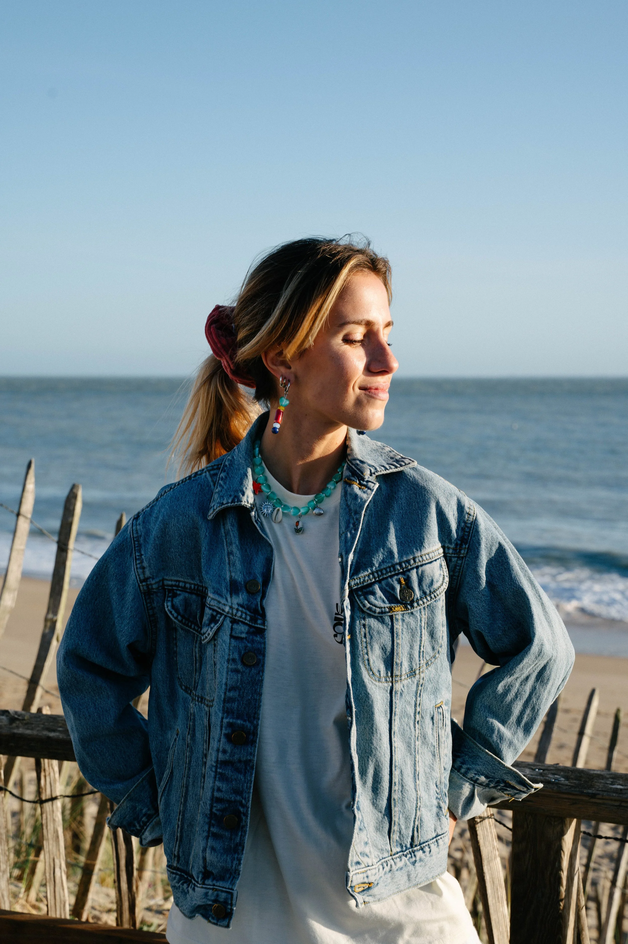 Femme portant une veste en jean et un t-shirt sur une plage, avec l'océan en arrière-plan.