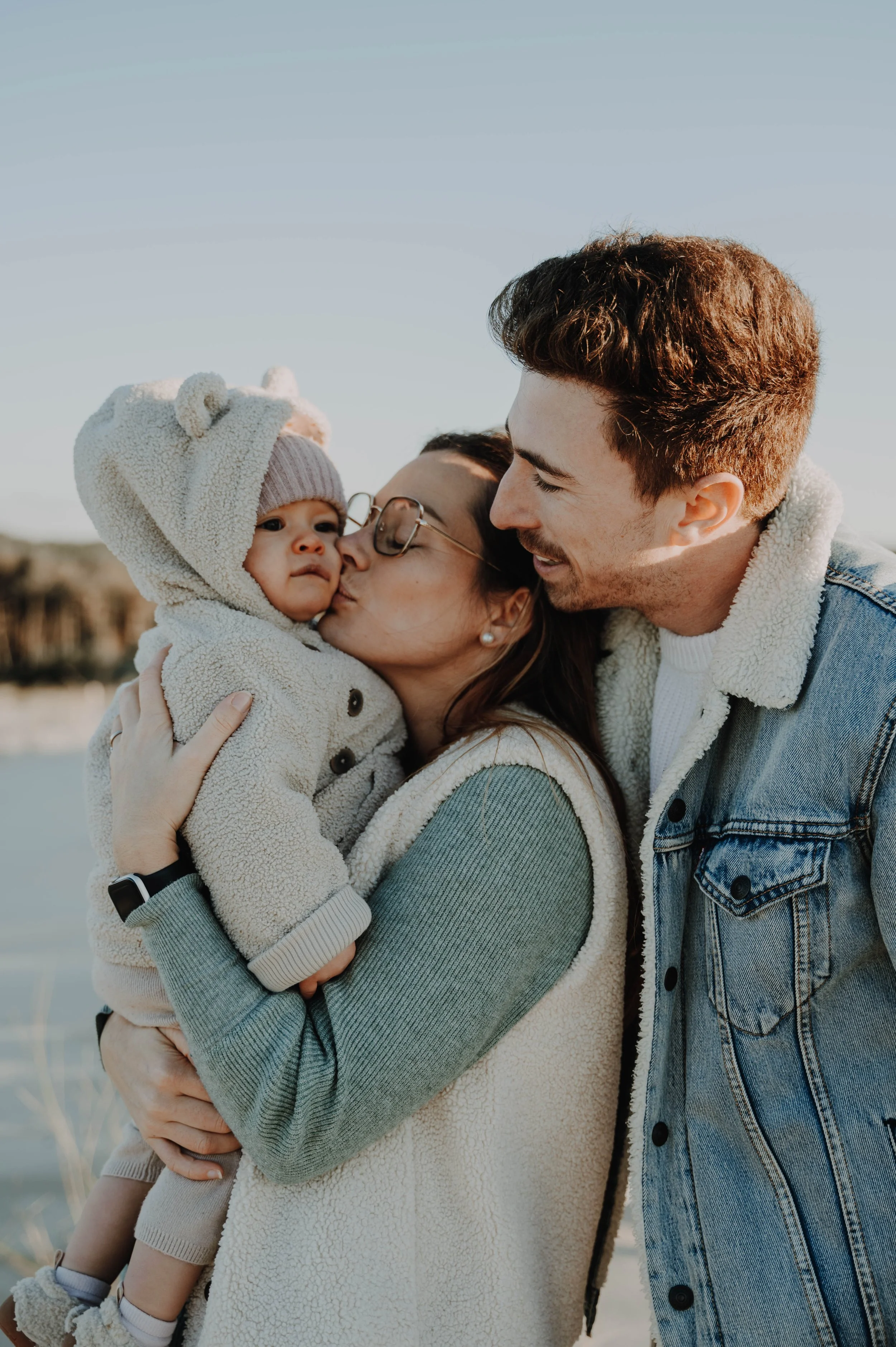 Deux adultes souriants, un homme et une femme, tenant et embrassant un bébé habillé chaudement, à l'extérieur, sous un ciel bleu.