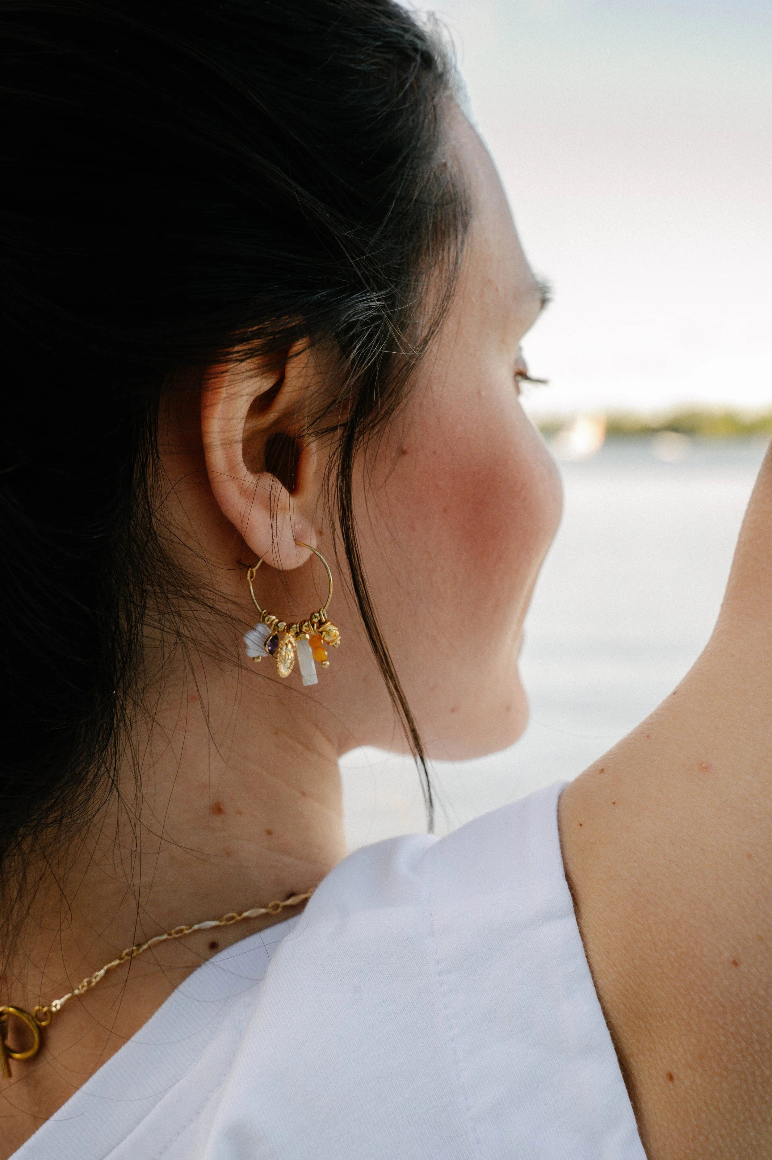Femme portant des boucles d'oreilles dorées avec des pendentifs colorés, vue de profil, fond flou.