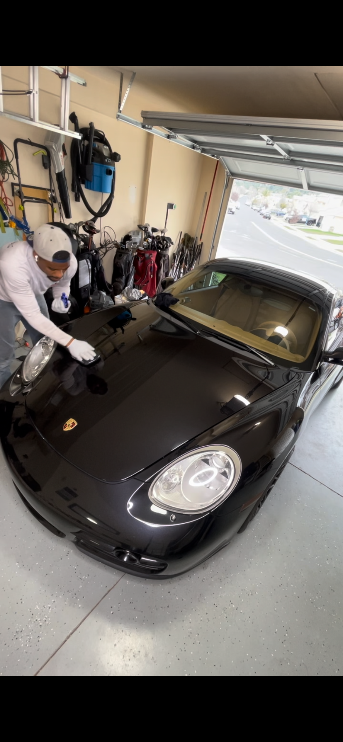 A person cleaning a black Porsche sports car inside a garage with tools and golf bags visible in the background.