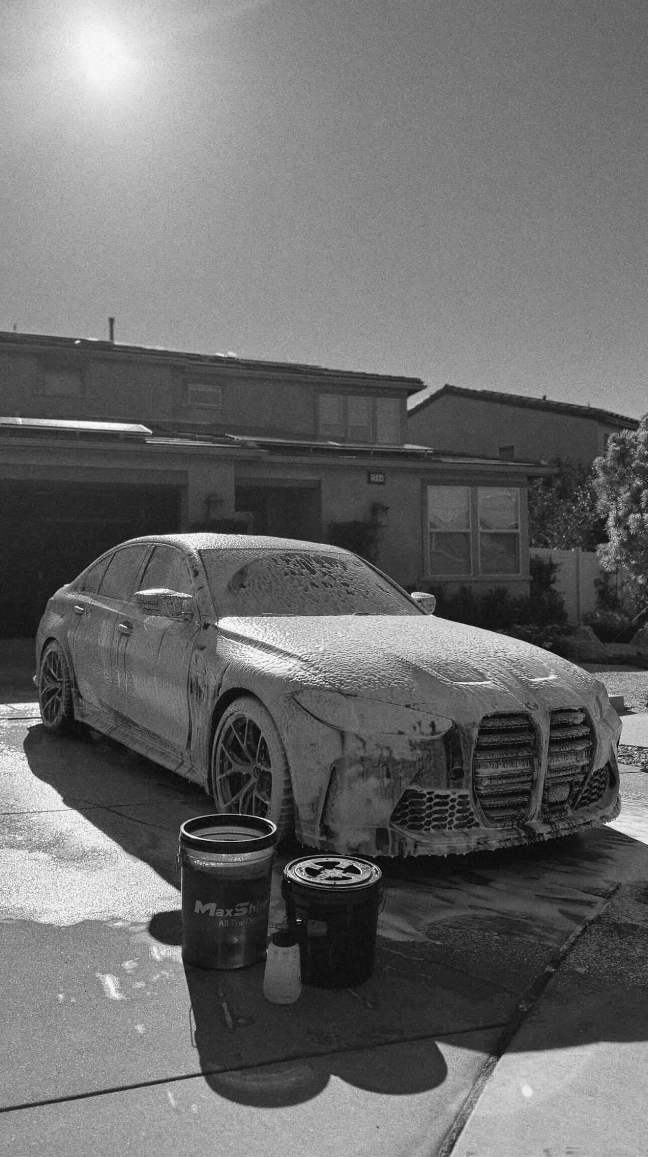 A car covered in soap suds and foam during a car wash on a driveway. Two buckets are on the ground in front of the car, with one bearing the MaxSh logo. The background shows a suburban house and a tree, with sunlight shining brightly.