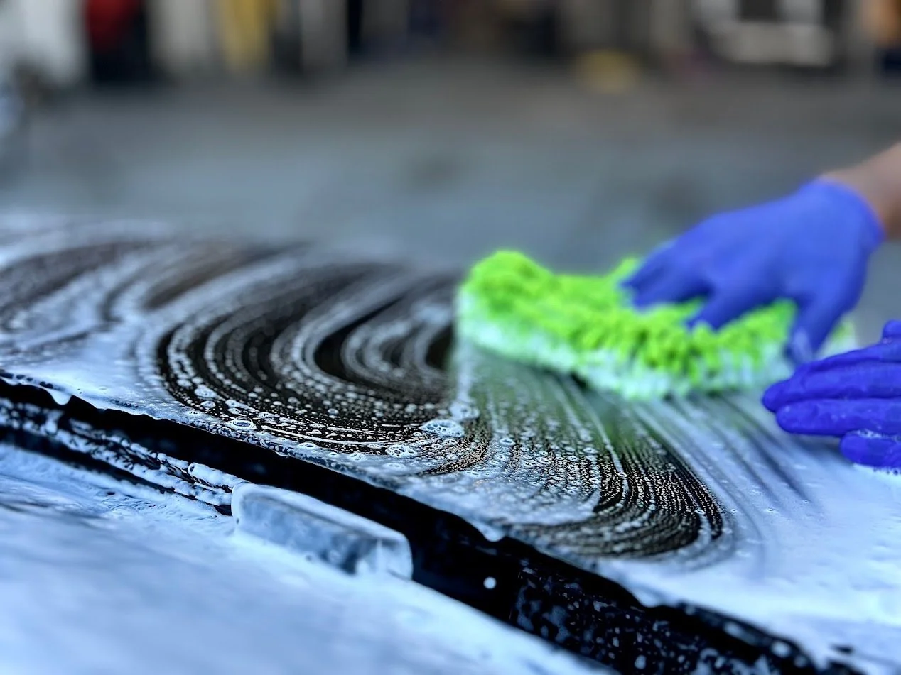 A person wearing blue gloves cleaning a soapy surface with a green scrub brush.