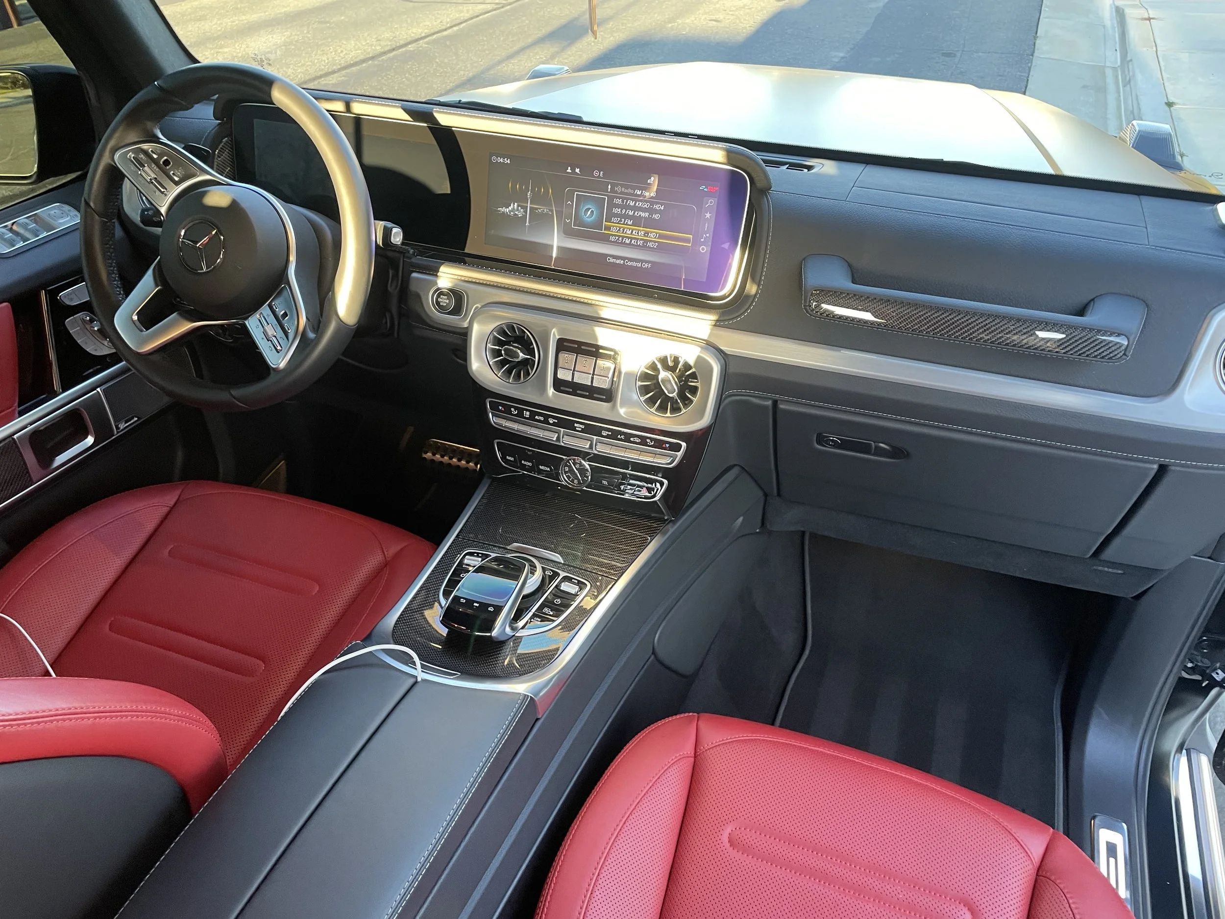 The interior of a vehicle with a black steering wheel, digital dashboard, and red leather seats. The dashboard features a large display screen showing radio station options. Outside the windshield, a driveway and a sidewalk are visible.