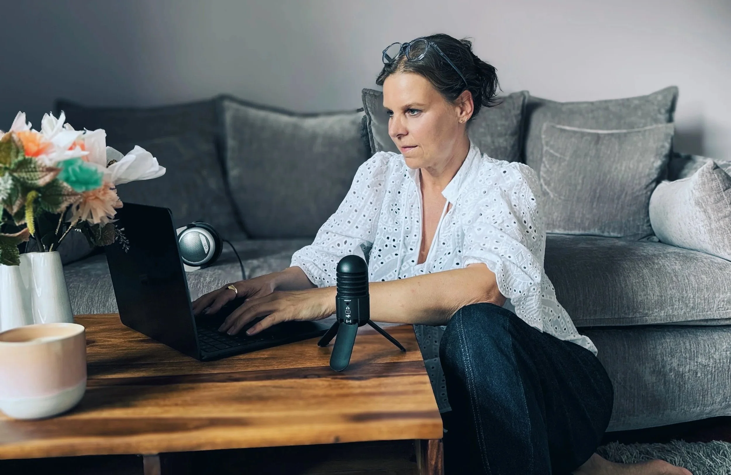 Podcast-Expertin Kathrin Pabst arbeitet an Laptop, sitzend auf dem Boden vor einem Sofa, mit Mikrofon und Kopfhörern, neben einem Kaffeebecher und Blumen auf einem Tisch.
