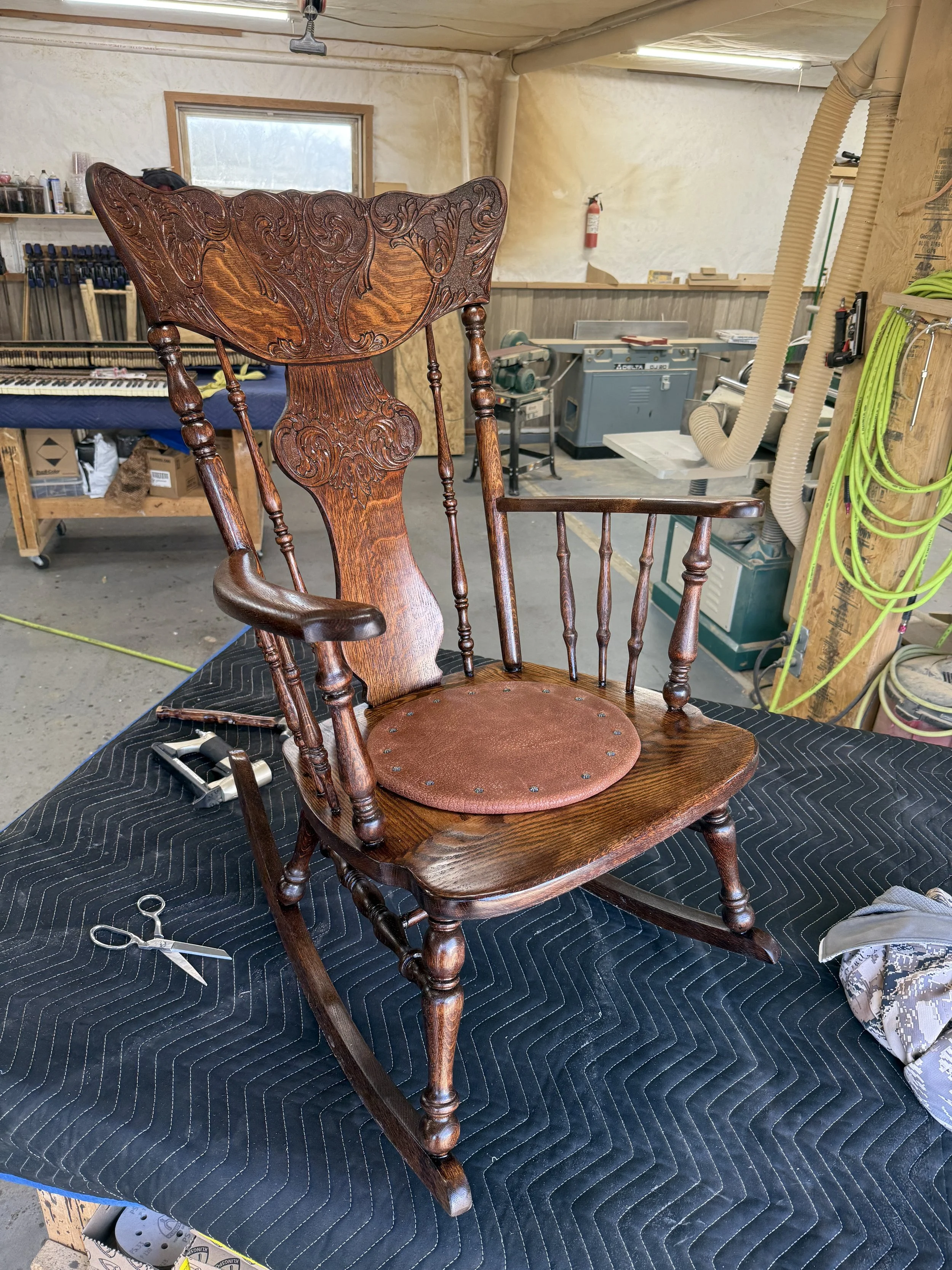 A wooden rocking chair with ornate carvings on the backrest and arms, placed on a black quilted surface in a woodworking workshop. Tools and equipment are visible in the background.