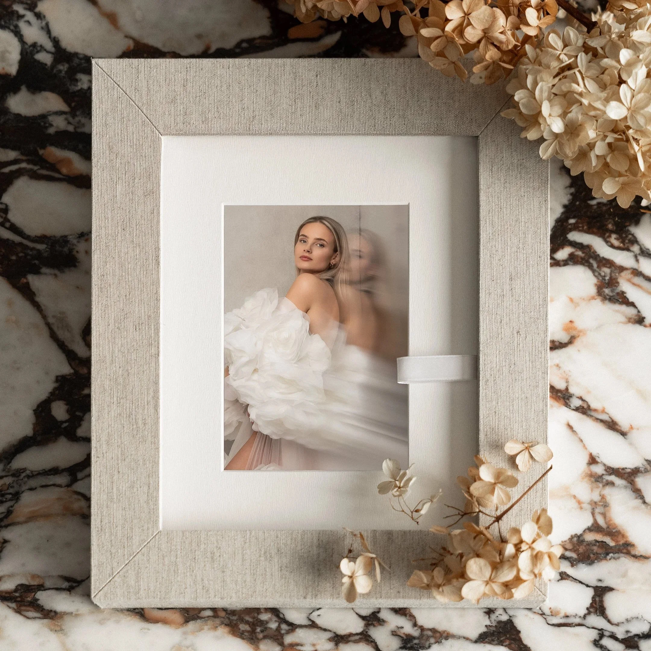 Fotografie eines jungen Frauenporträts in einem eleganten weißen Kleid, präsentiert in Rahmen auf einem Marmor-Tisch, geschmückt mit beige getrockneten Blumen.